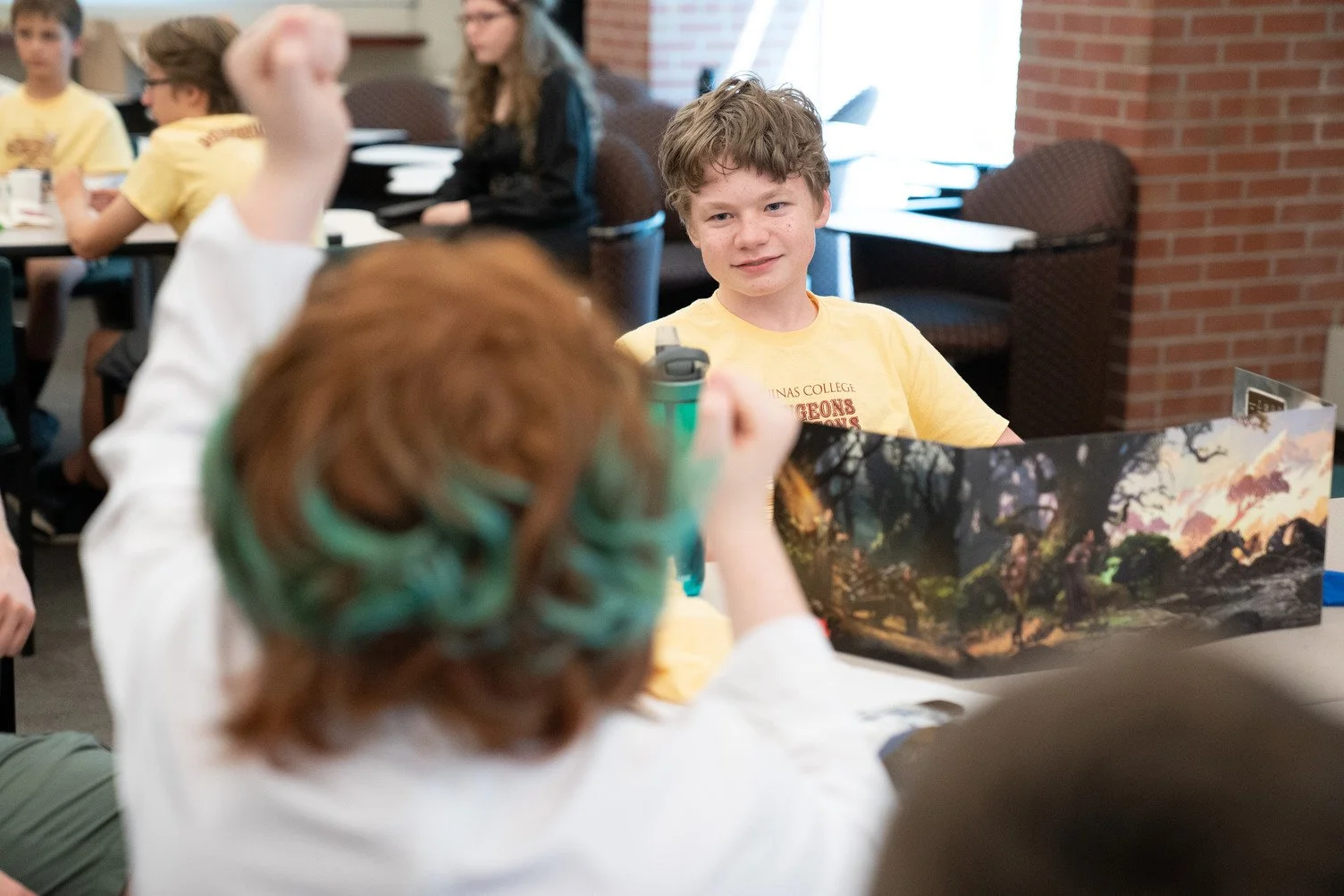 A classroom scene with students at tables, one boy in a yellow T-shirt looking at another student with green hair and a white shirt, who is holding a game or storybook with a fantasy landscape.