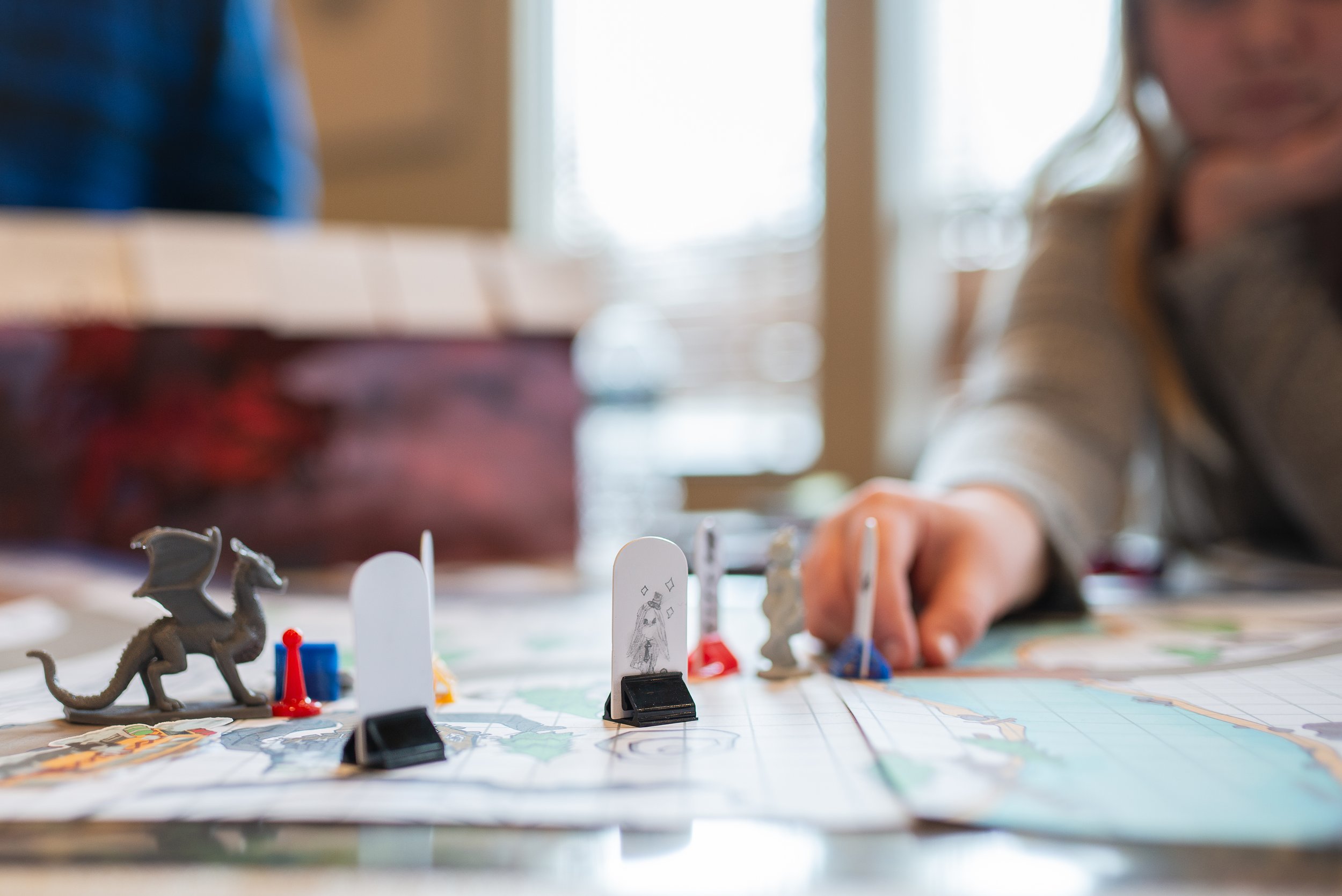 Close-up of a board game with fantasy-themed figurines and player hand reaching to make a move, set indoors with natural light.