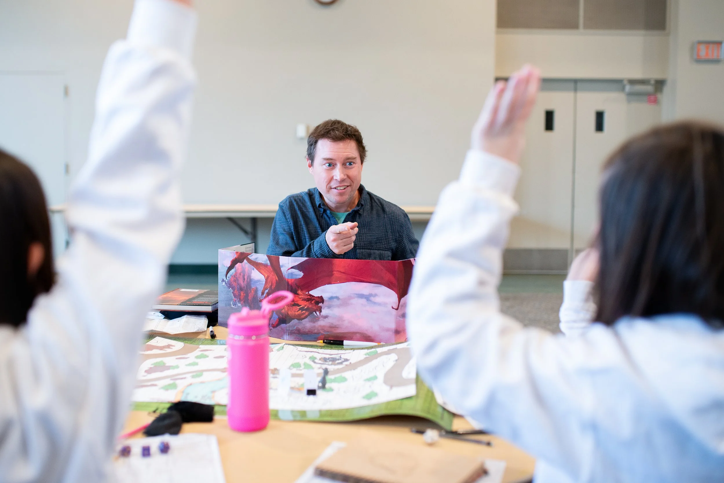 A person running a tabletop role-playing game sessions with children, using a dragon-themed screen, a map, and miniatures, with children raising their hands.