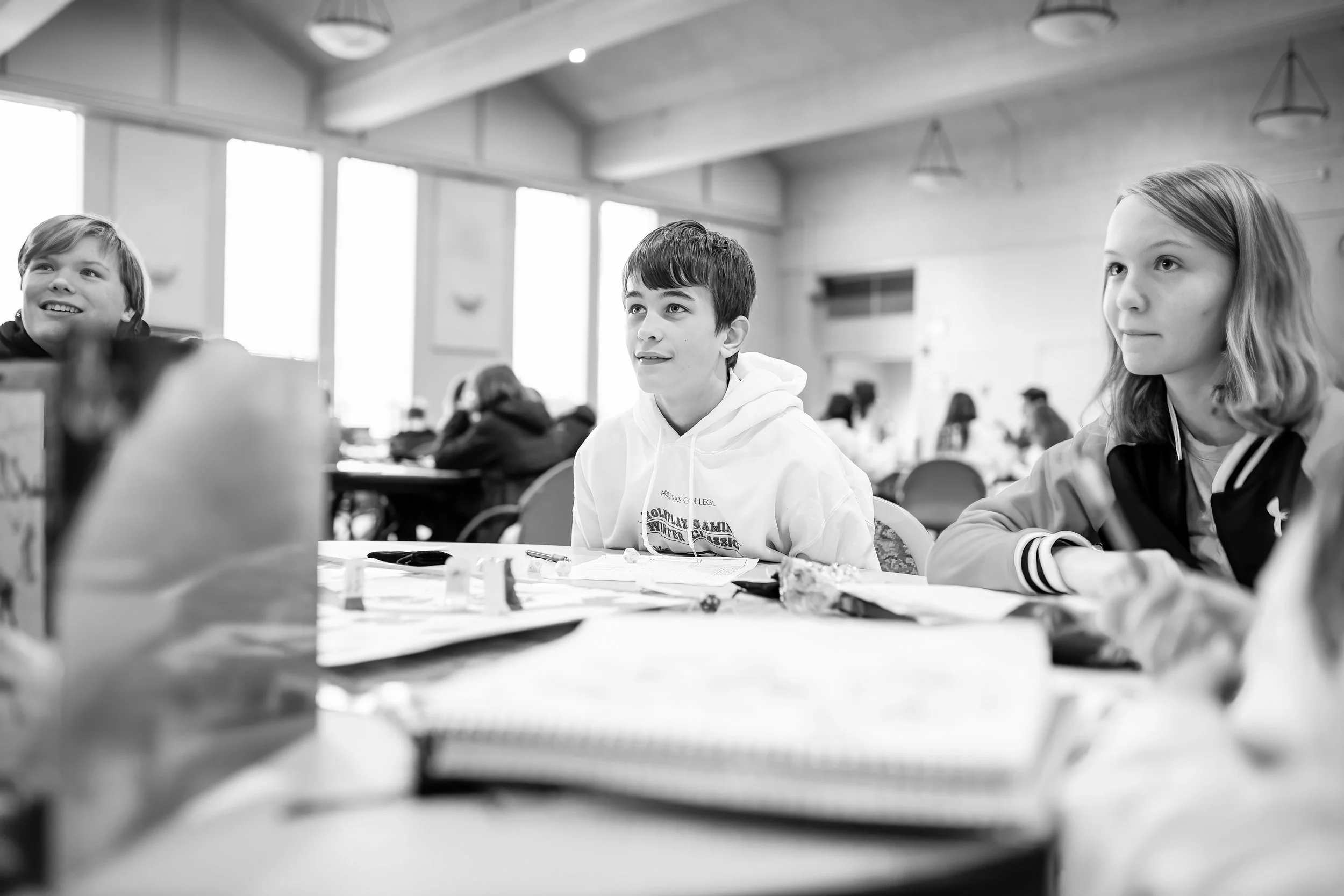 Black and white photo of children sitting at a table in a bright room, listening attentively, with other people in the background.