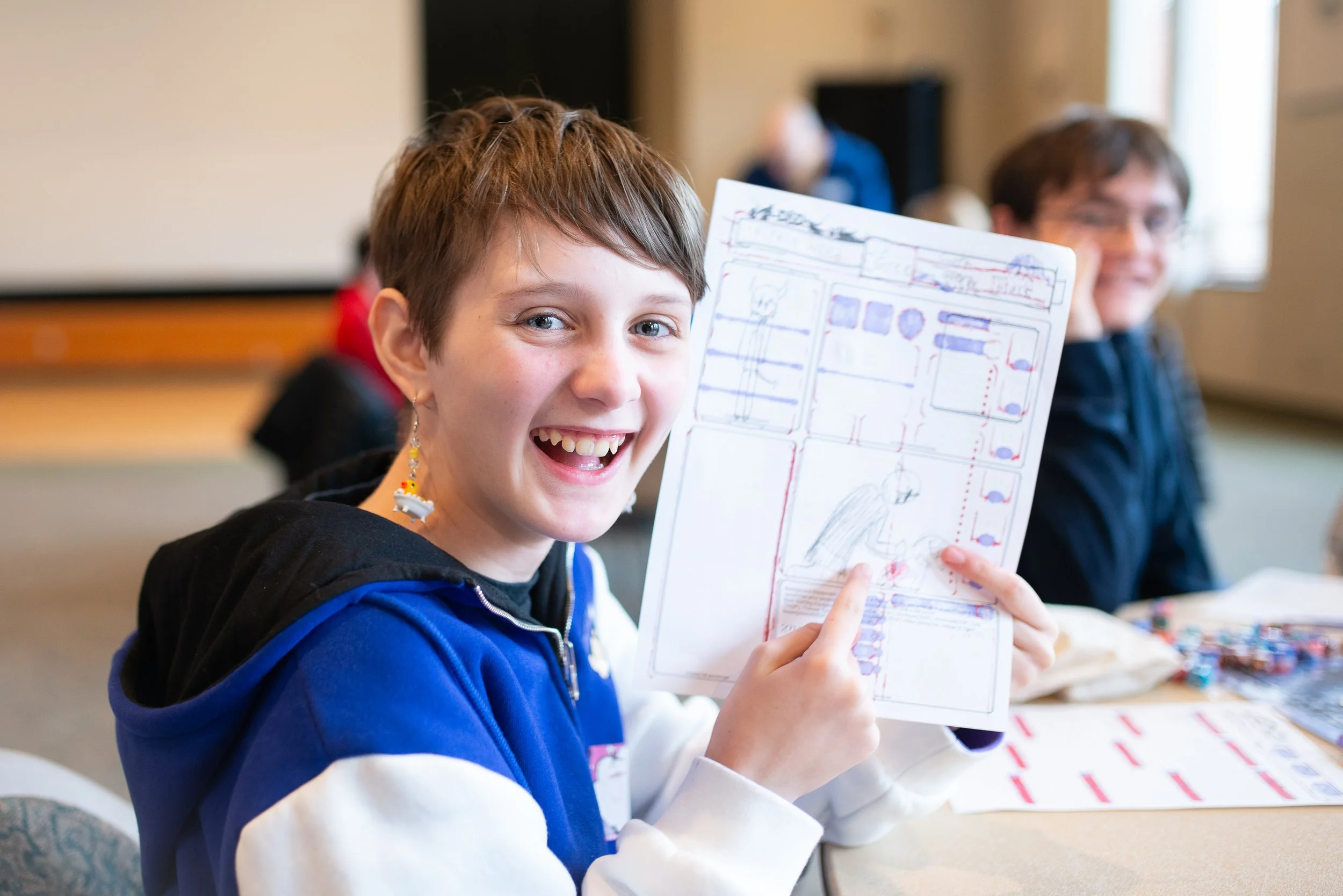A young girl with short brown hair and earrings smiling while holding up a comic book or graphic novel in a classroom or classroom-like setting.