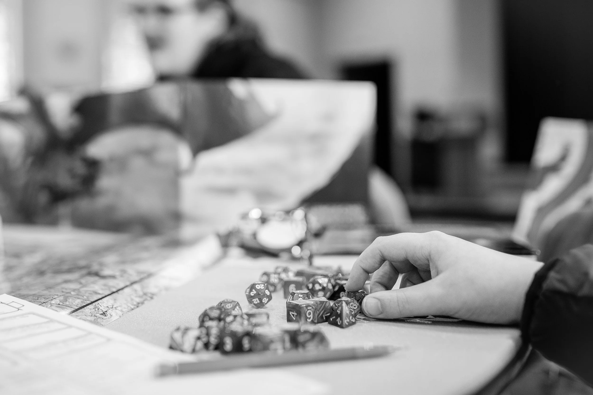 Black and white photo of a person rolling dice on a tabletop, with a map, a pen, and other gaming accessories visible.