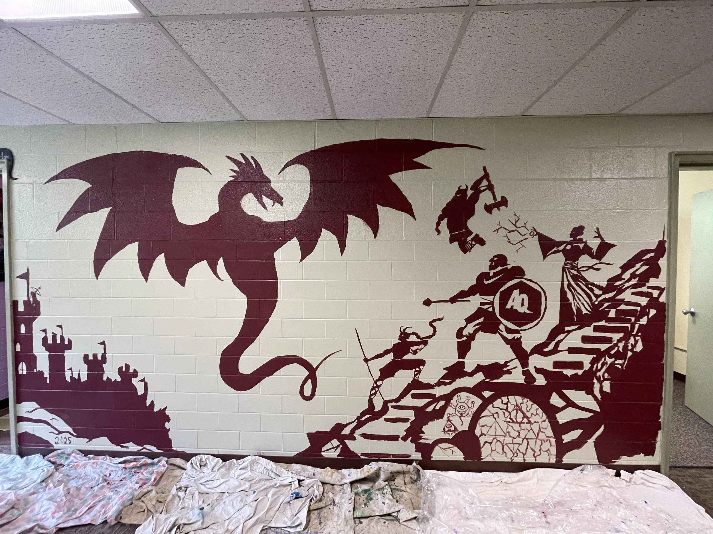 A large painted mural on a wall depicts a dragon with large wings and a long tail, along with a group of medieval warriors fighting on a staircase, some holding shields and weapons. The mural is painted in dark red against a light-colored brick wall.