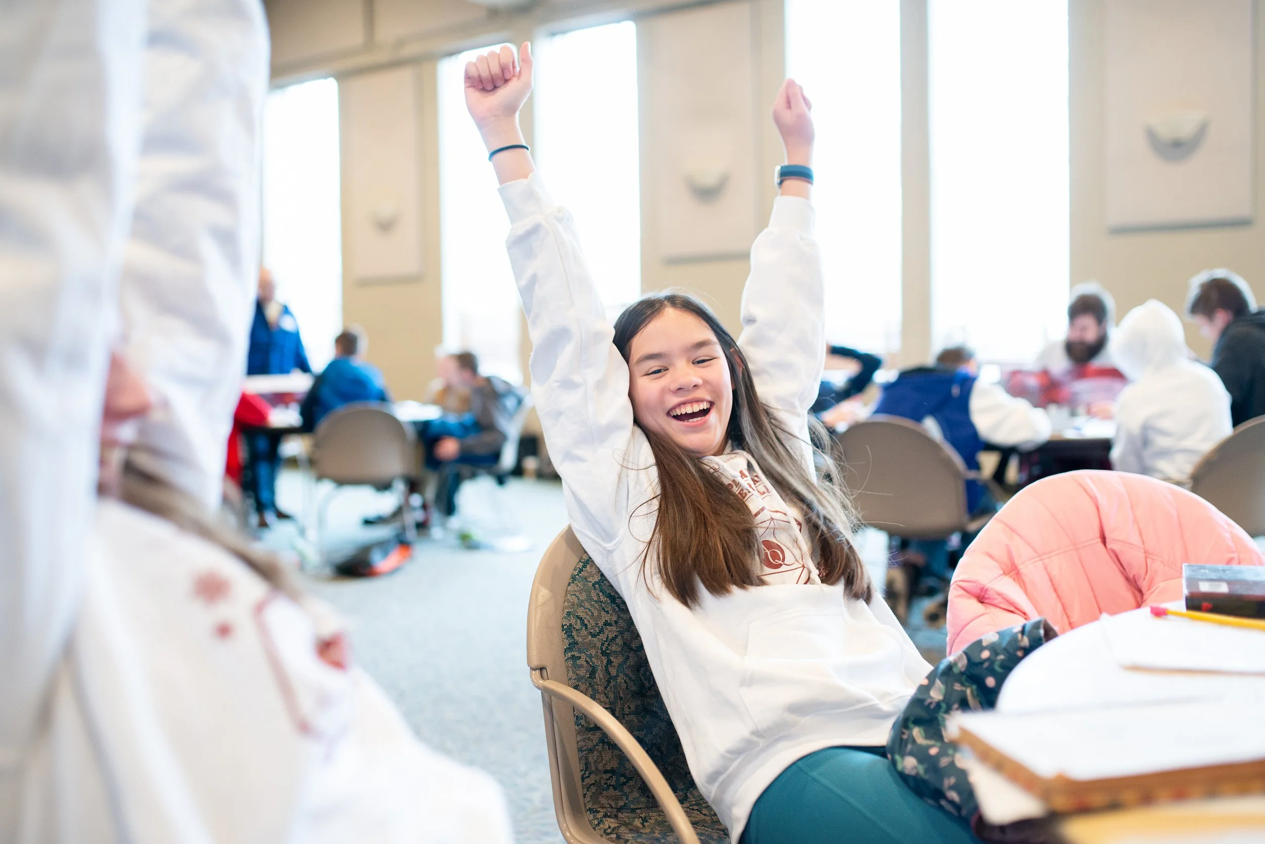 A young girl with long brown hair, wearing a white long-sleeve shirt, is sitting at a table with her arms raised in victory or excitement. She is smiling broadly in a lively indoor setting with other children and adults in the background.