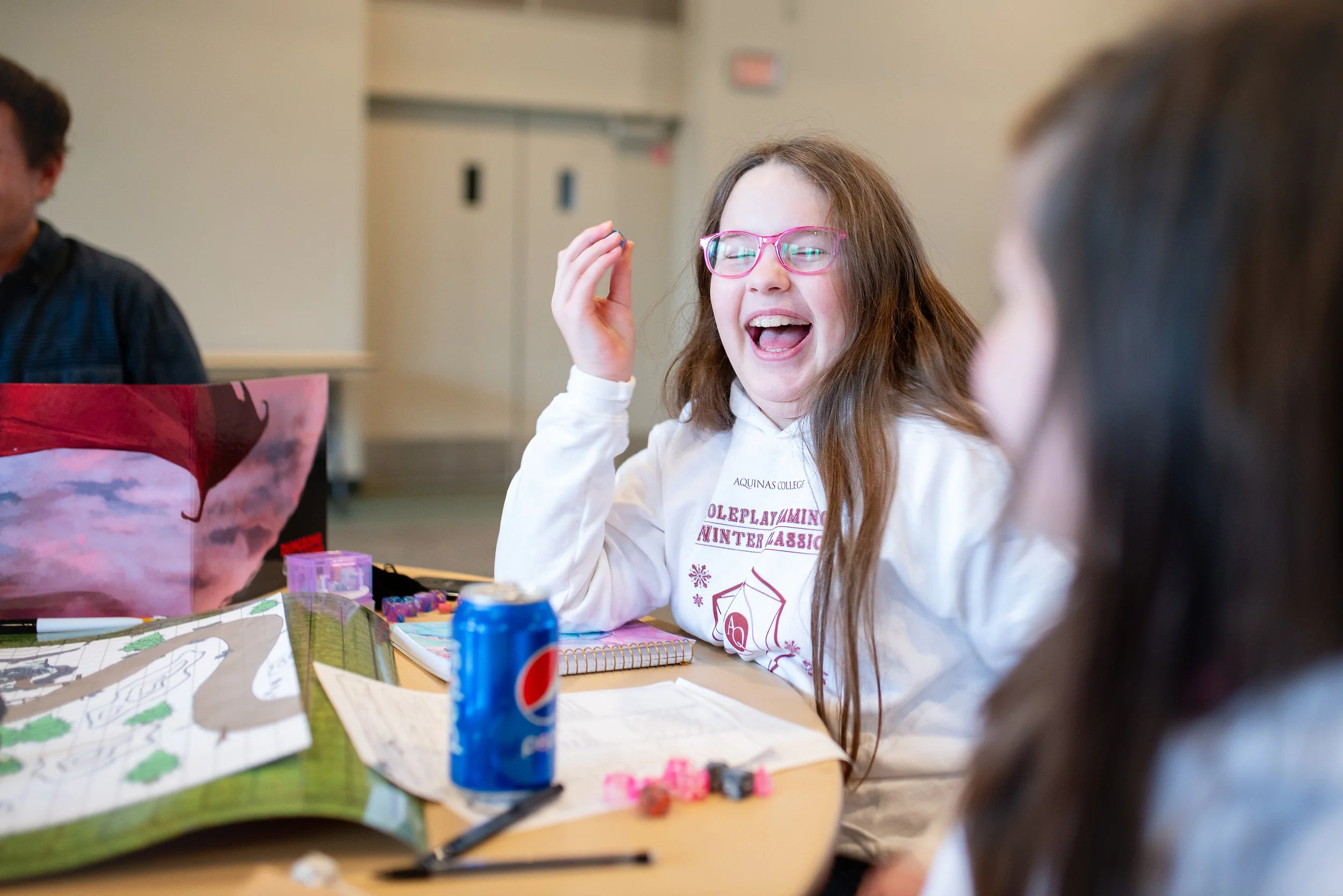 A girl with long brown hair, pink glasses, and a white sweatshirt smiling and laughing during a social gathering at a table with snacks, a Pepsi can, and a game board. She appears to be having fun.