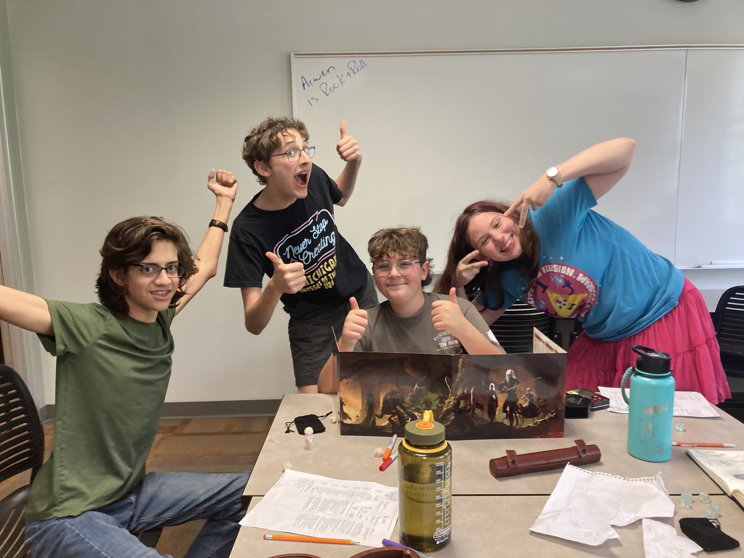 Four young people are gathered around a table with a fantasy tabletop game, smiling and giving thumbs up. A whiteboard behind them has writing that says, "Auren is Rude!!" There are water bottles, papers, and game accessories on the table.