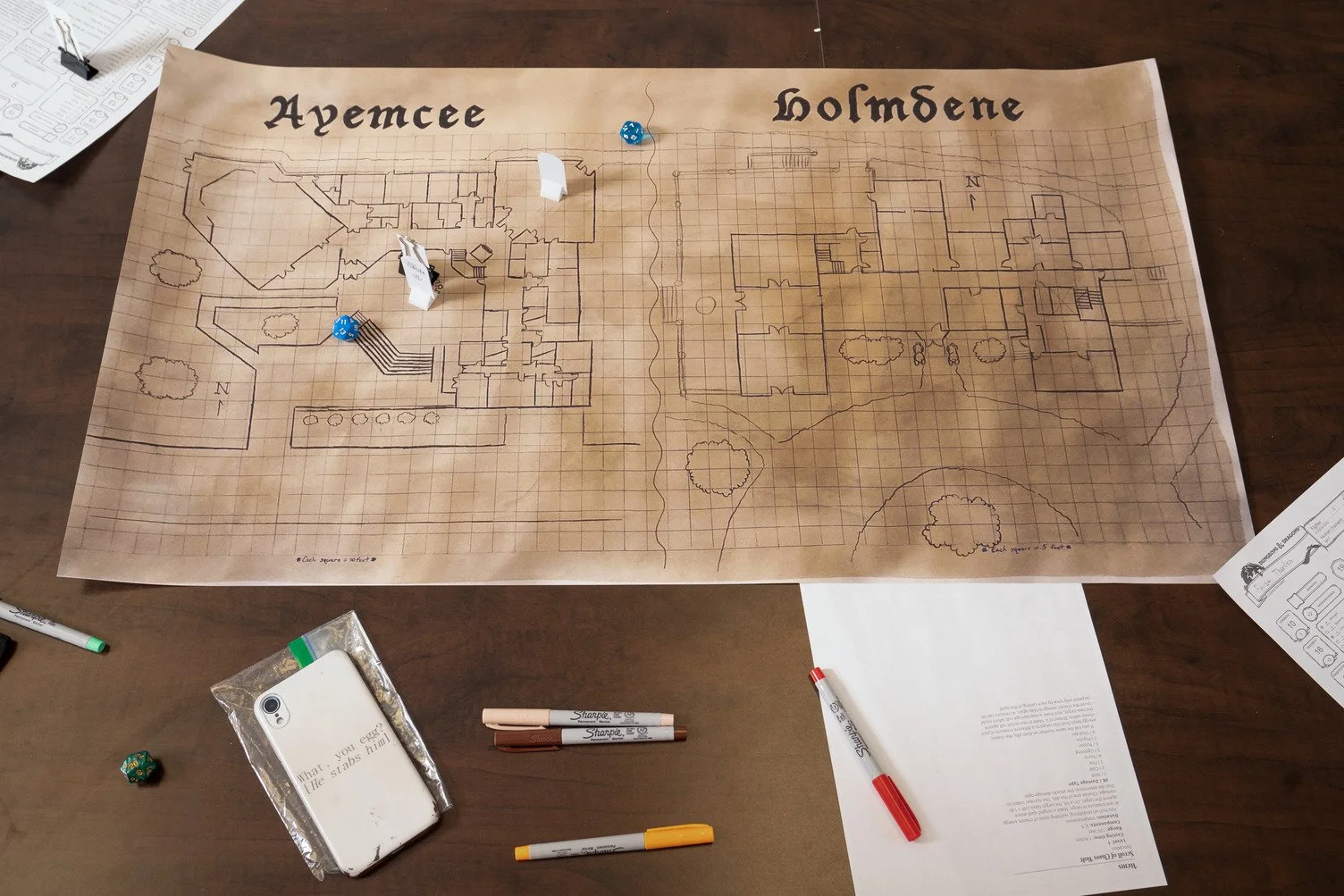 Table with a large hand-drawn diagram of a house layout, labeled 'Avenue' and 'Homdene.' There are game pieces including white and blue markers and dice on the diagram, suggesting a game in progress. Surrounding the diagram are various markers, pens,