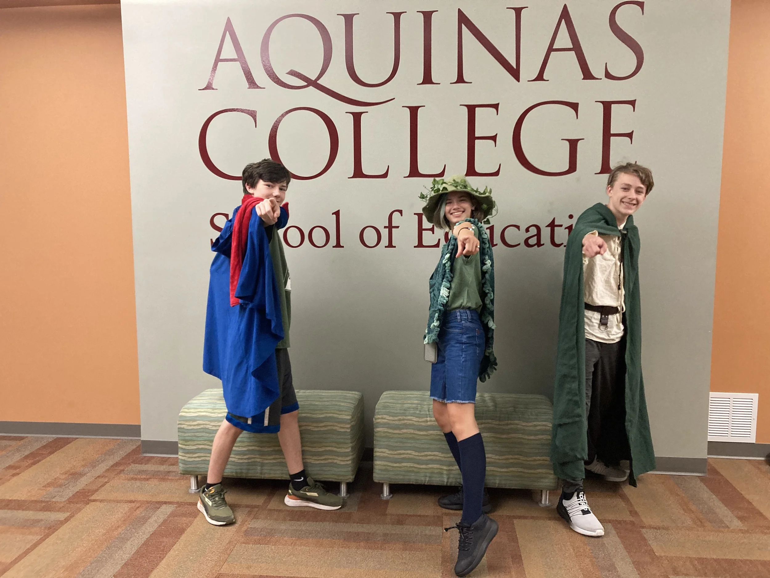 Three students dressed as superheroes and characters, standing in front of a sign that reads 'Aquinas College School of Education.' They are pointing towards the camera, with two standing on a green bench and one in front of it.