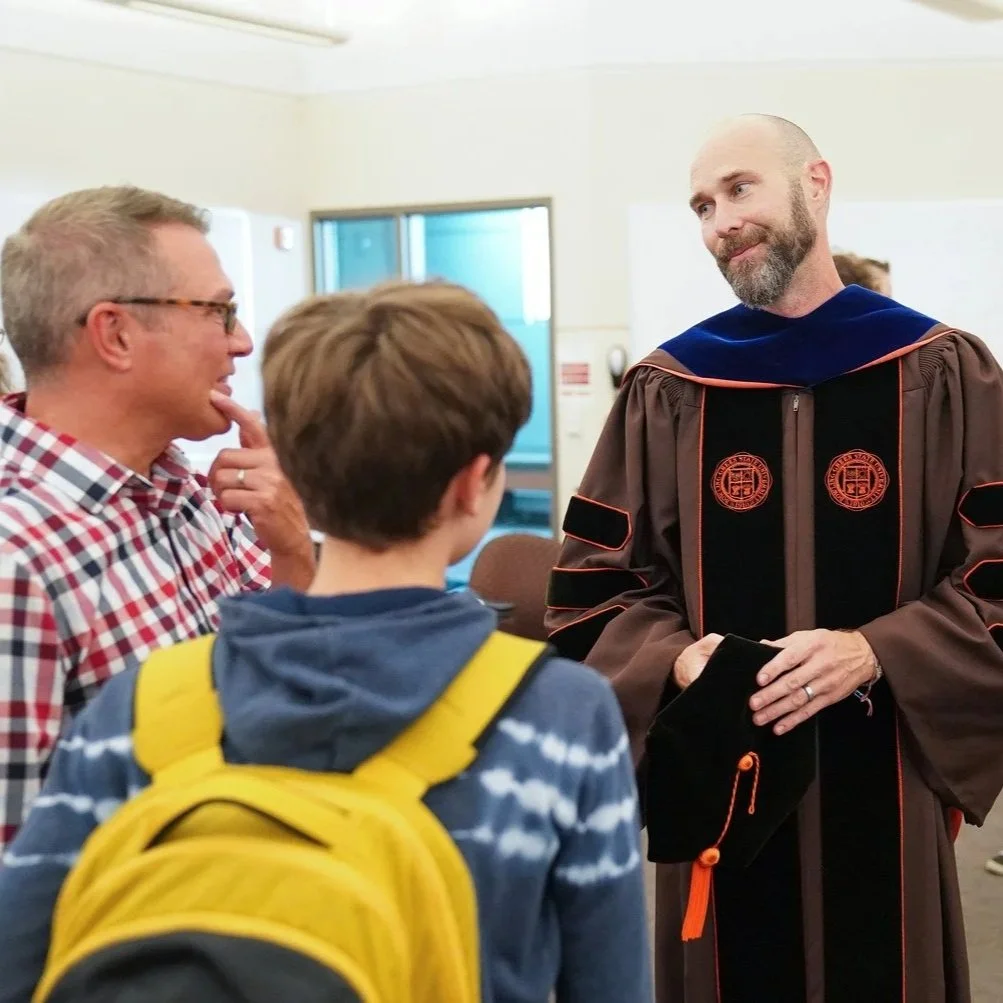 People talking, including a graduate in robes, in an indoor setting.