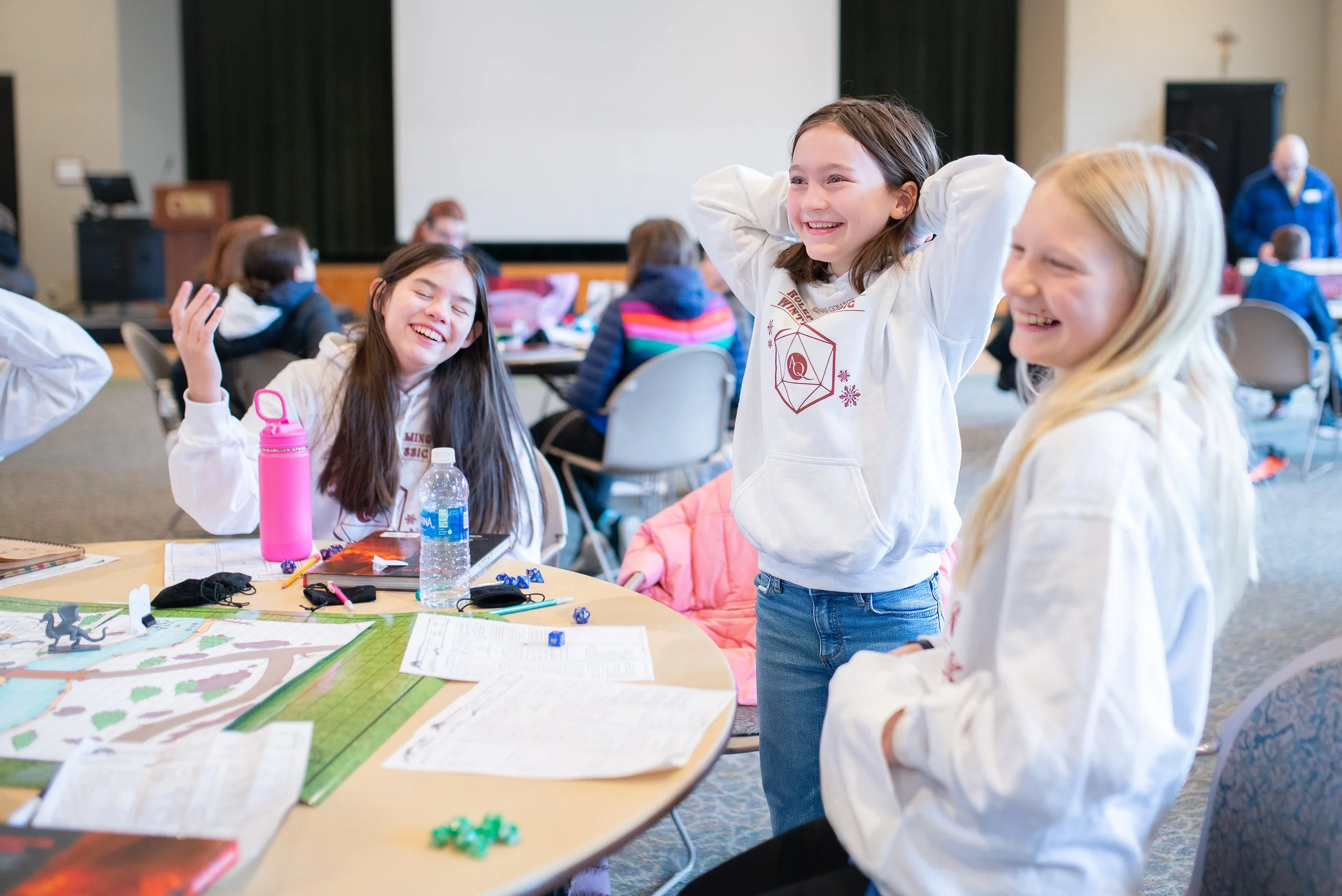 Three young girls laughing and playing at a table with game pieces and papers, in a crowded indoor event space.