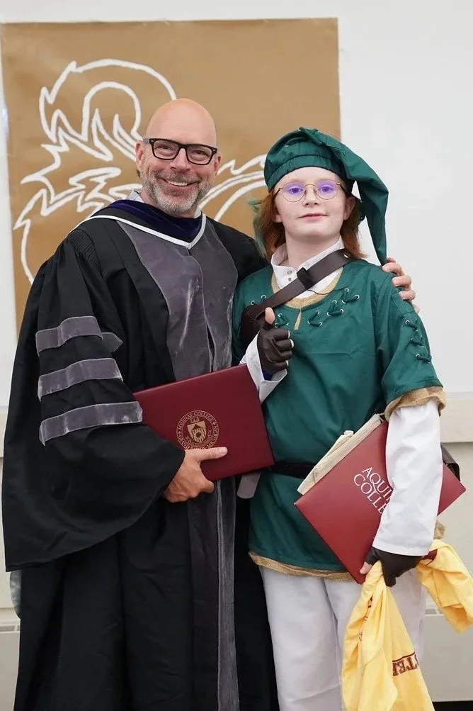 Two individuals in academic regalia standing together, one adult man and one young person, at a graduation or academic event. The adult is wearing a black gown with velvet panels and stripes on the sleeves, holding a red diploma folder. The young person is dressed in a green and gold costume with a matching hat, glasses, and gloves, also holding diploma folders and a yellow cloth.