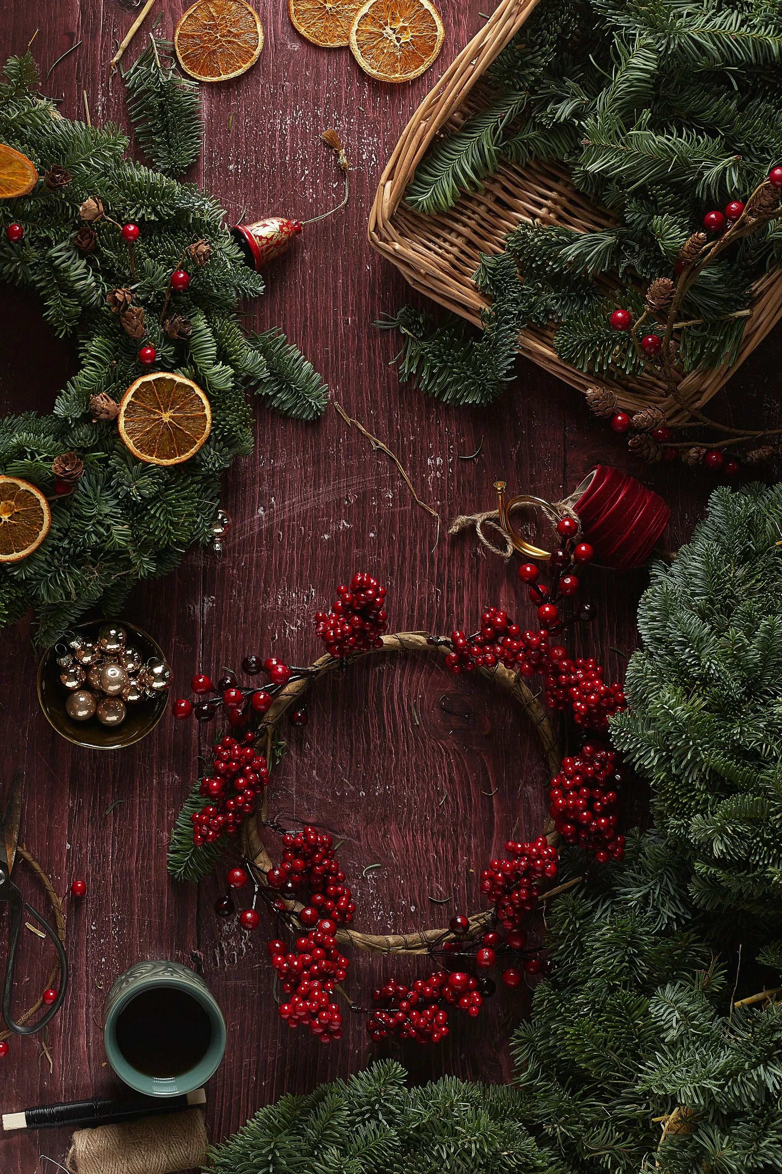 Flat lay of Christmas wreath-making supplies on a wooden surface, including evergreen branches, dried orange slices, red berries, ornaments, scissors, and a mug.