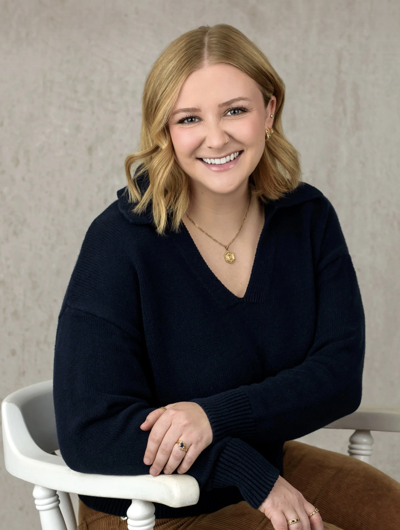 Young woman with blonde hair, wearing a black V-neck sweater, seated on a white chair, smiling, wearing gold jewelry including necklace, rings, earrings, and nose ring, against a textured gray background.