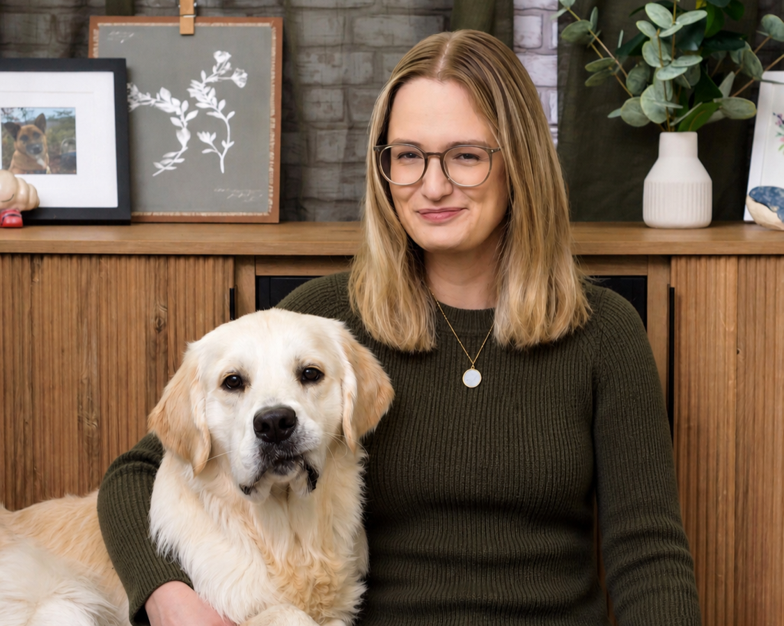 A woman with glasses and shoulder-length blonde hair sitting indoors with a golden retriever dog on her lap, in front of a wooden cabinet with framed pictures, artwork, a potted plant, and decorative items.