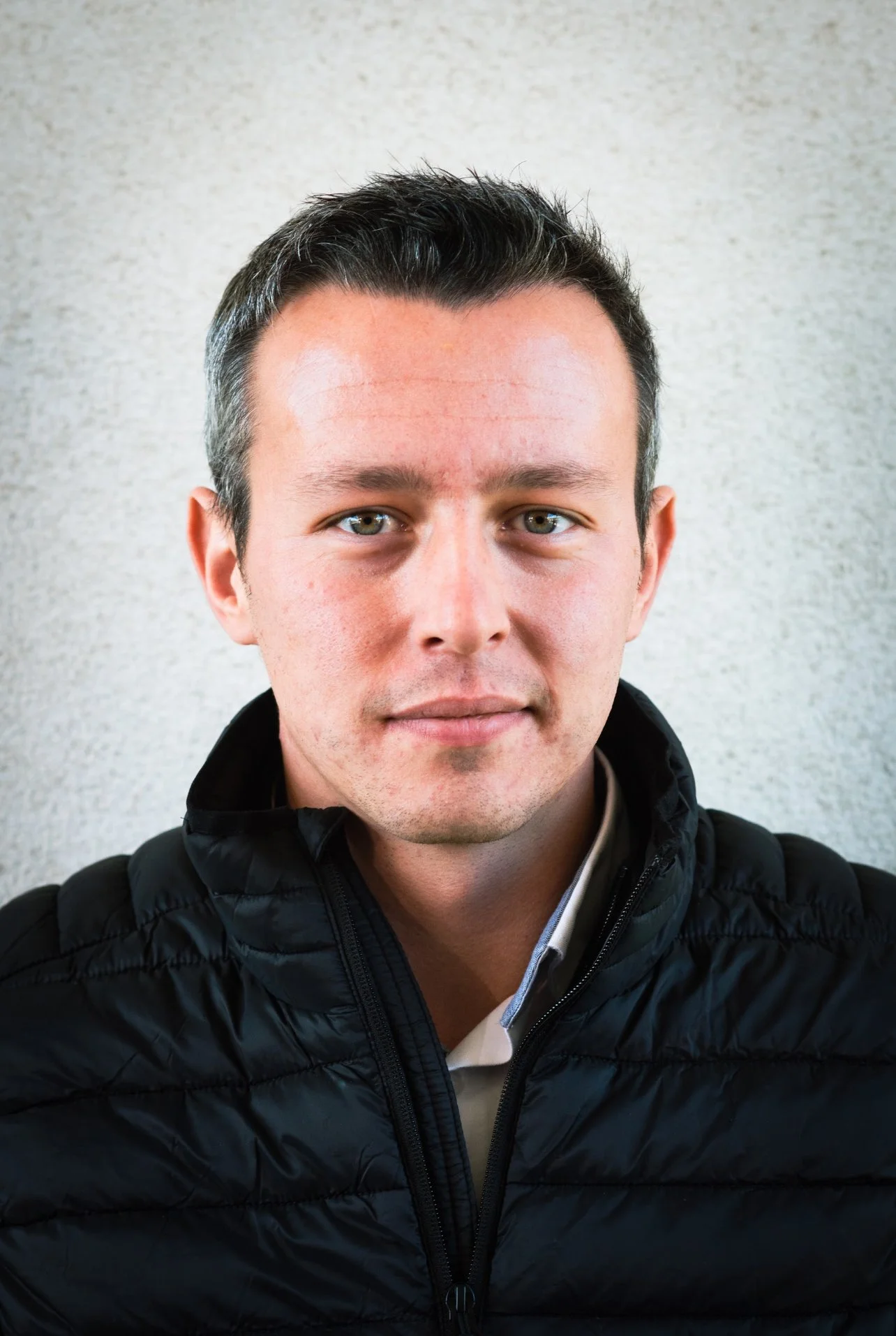 Portrait of a man with short dark hair, light skin, and blue eyes, wearing a black puffer jacket and a white shirt, standing against a textured gray background.