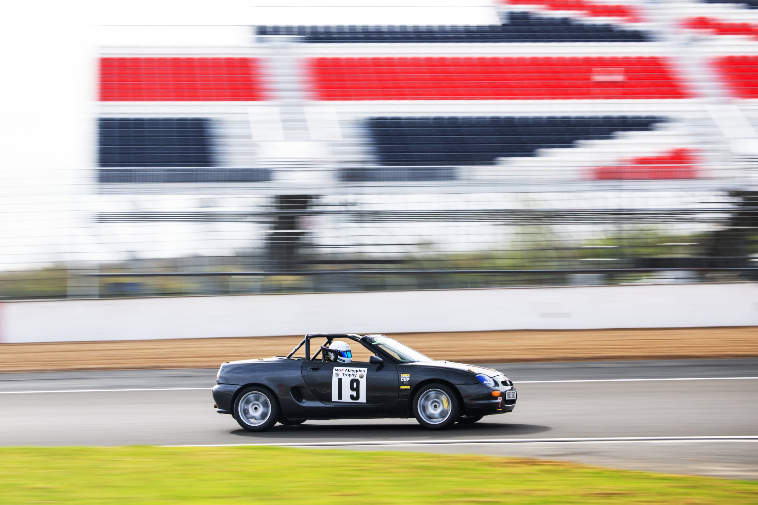 A black race car with the number 19 on its side, speeding on a race track with blurred background of grandstands and fencing.