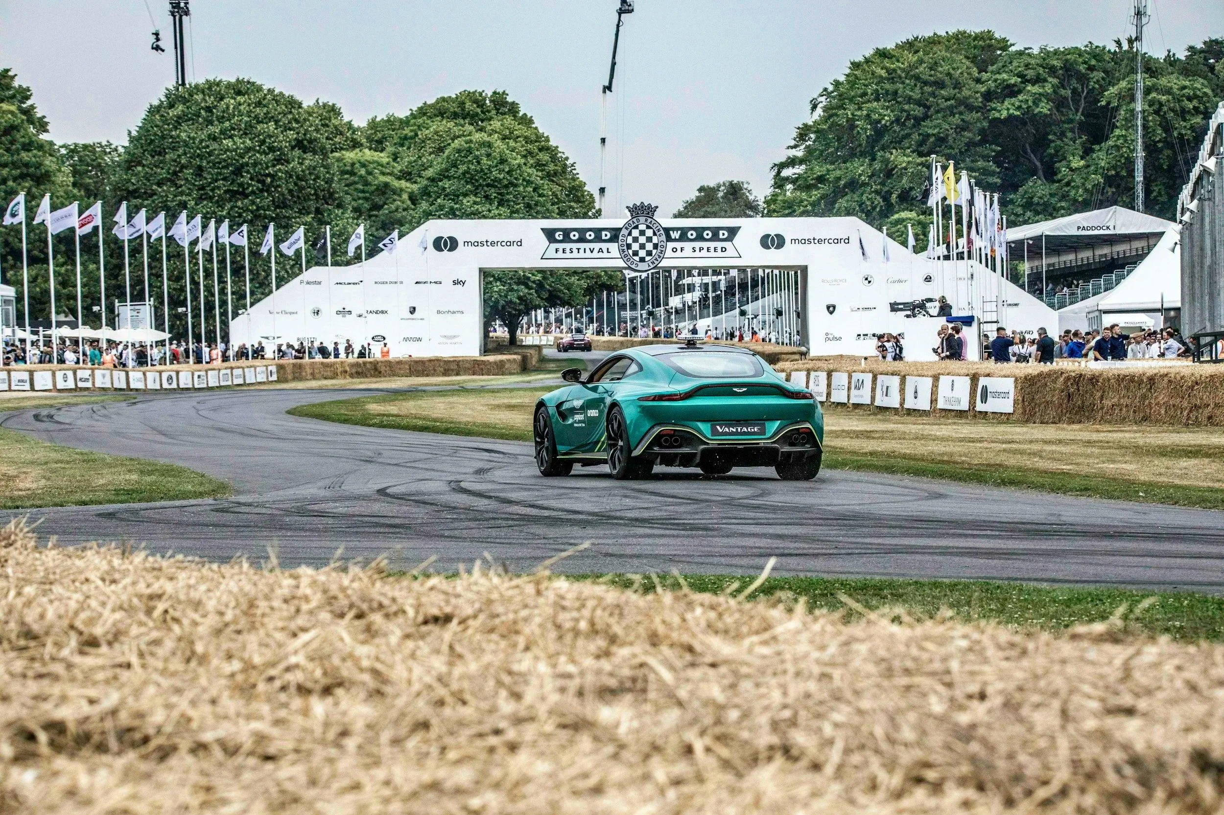A green Aston Martin Vantage race car on a track at the Goodwood Festival of Speed, with a large white festival arch and trees in the background, and a crowd of spectators along the sides.