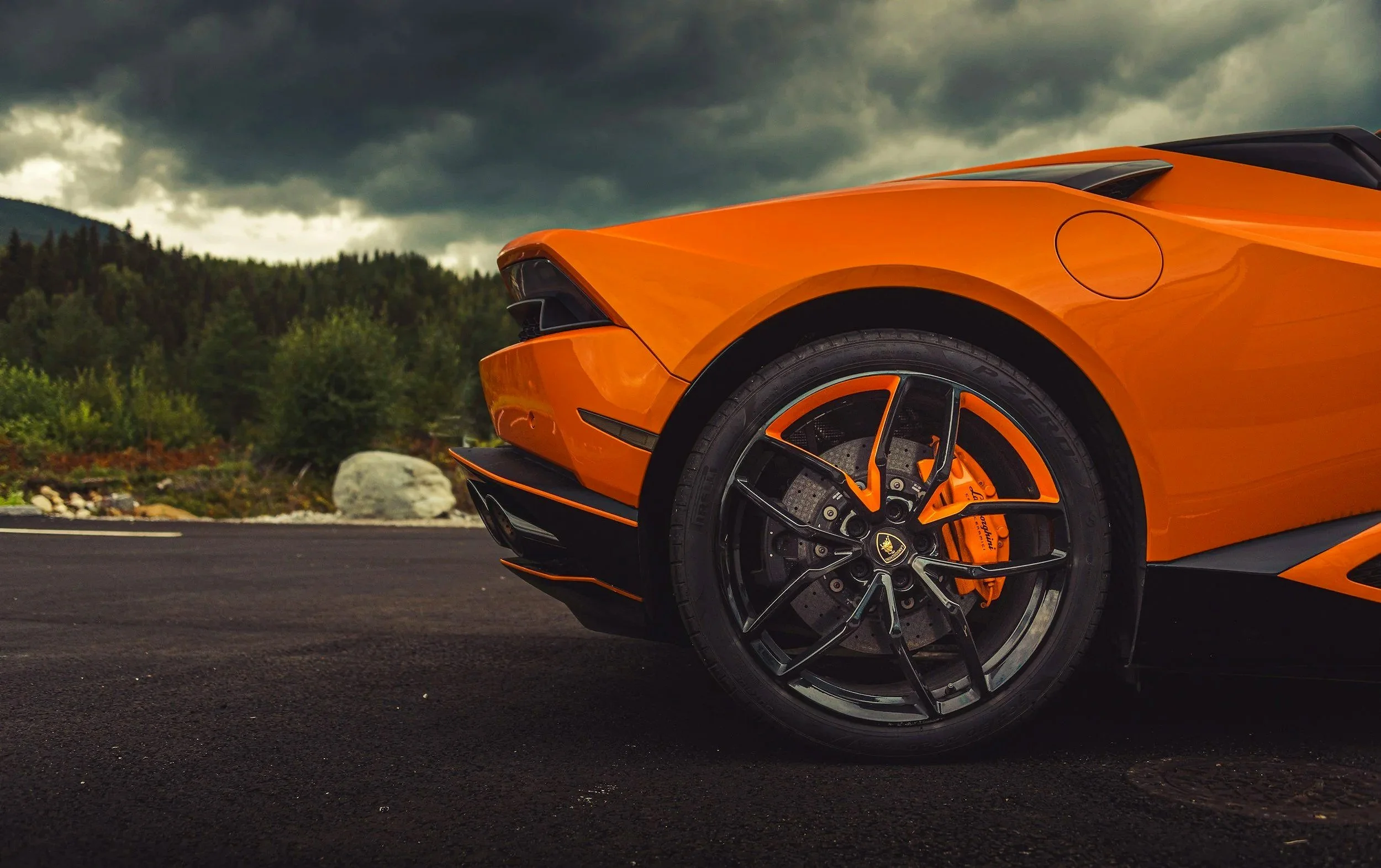Close-up of a bright orange sports car parked on a road with a scenic background of green trees and cloudy sky.