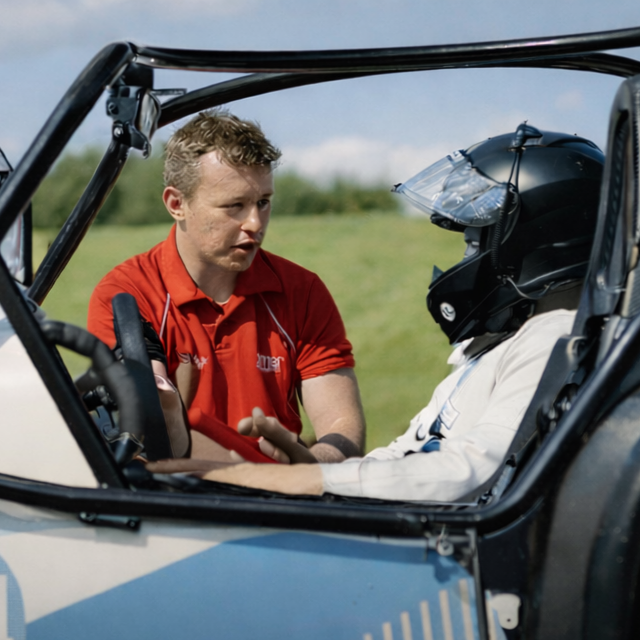 A man in a red shirt speaking to a person in a racing helmet inside a small open-cockpit racing car on a grassy field.