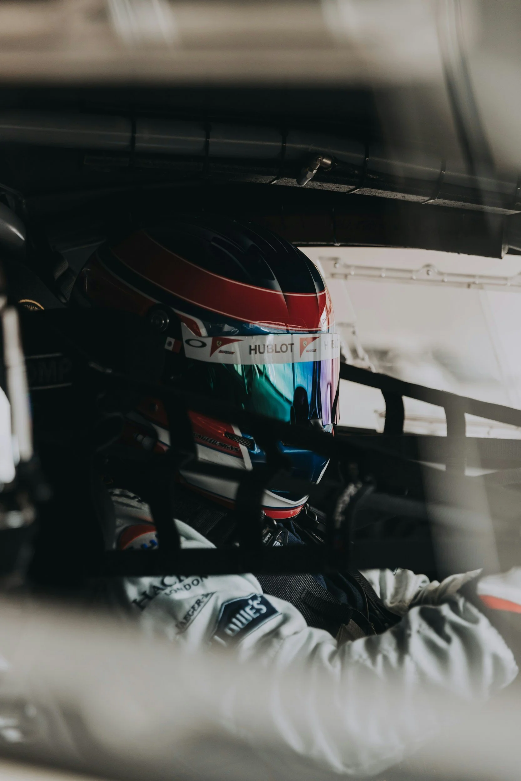 Close-up of a race car driver wearing a white racing suit and a red, black, and blue helmet with the Hublot logo, seated in a race car.