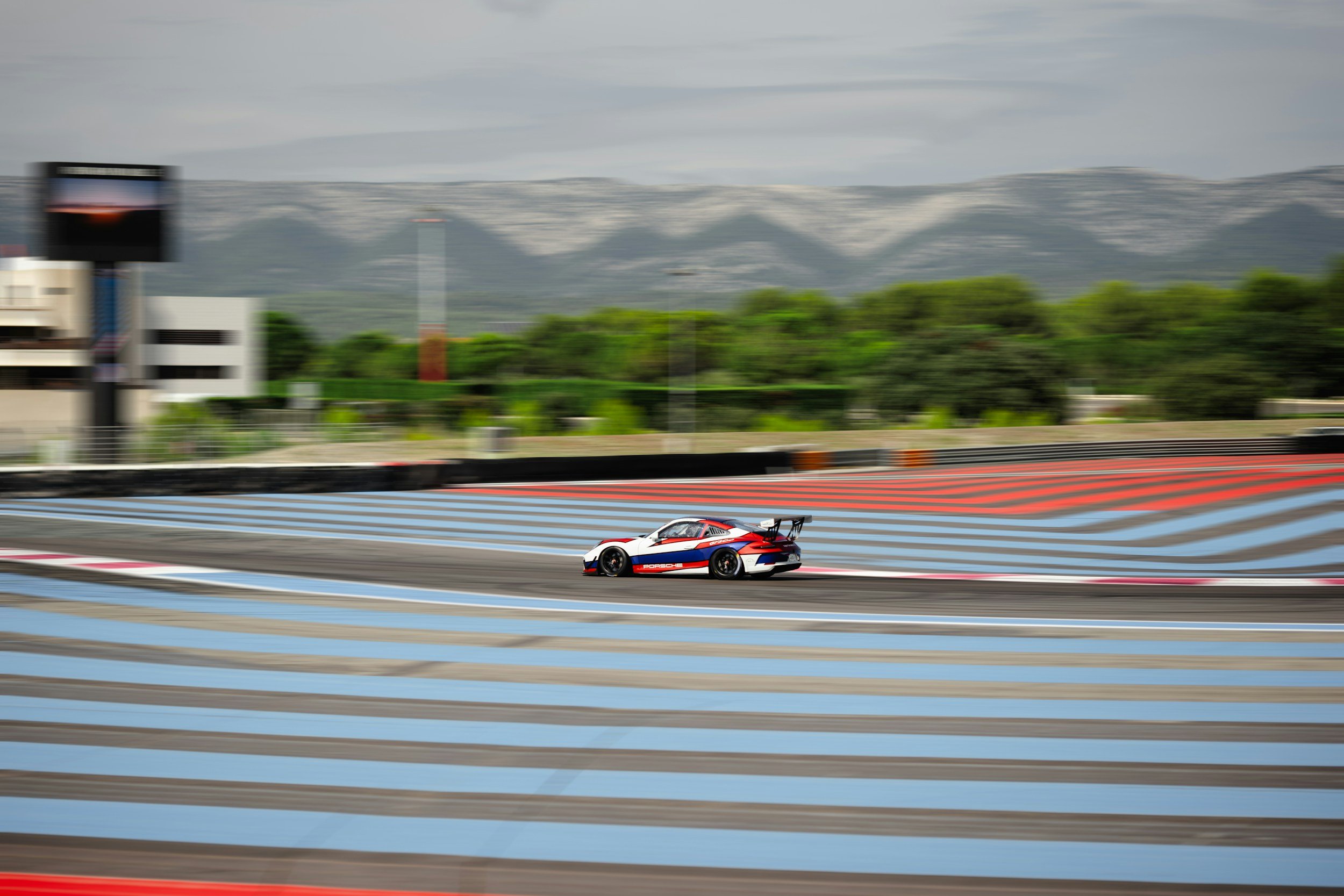 A race car on a racetrack with blue and red striped pavement, mountains in the background, and cloudy skies.