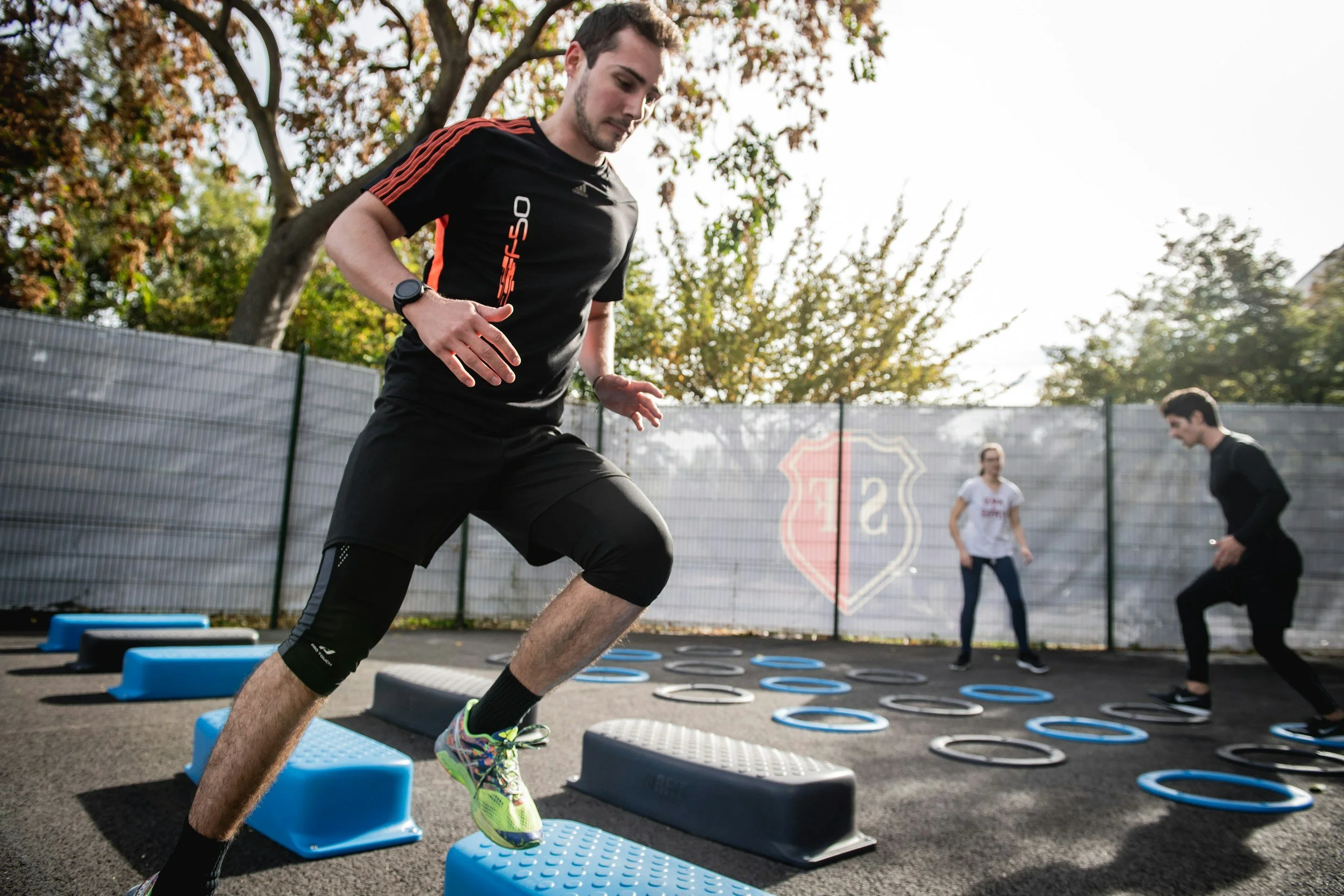 Man running and jumping over agility obstacles during outdoor fitness training with other people in the background.