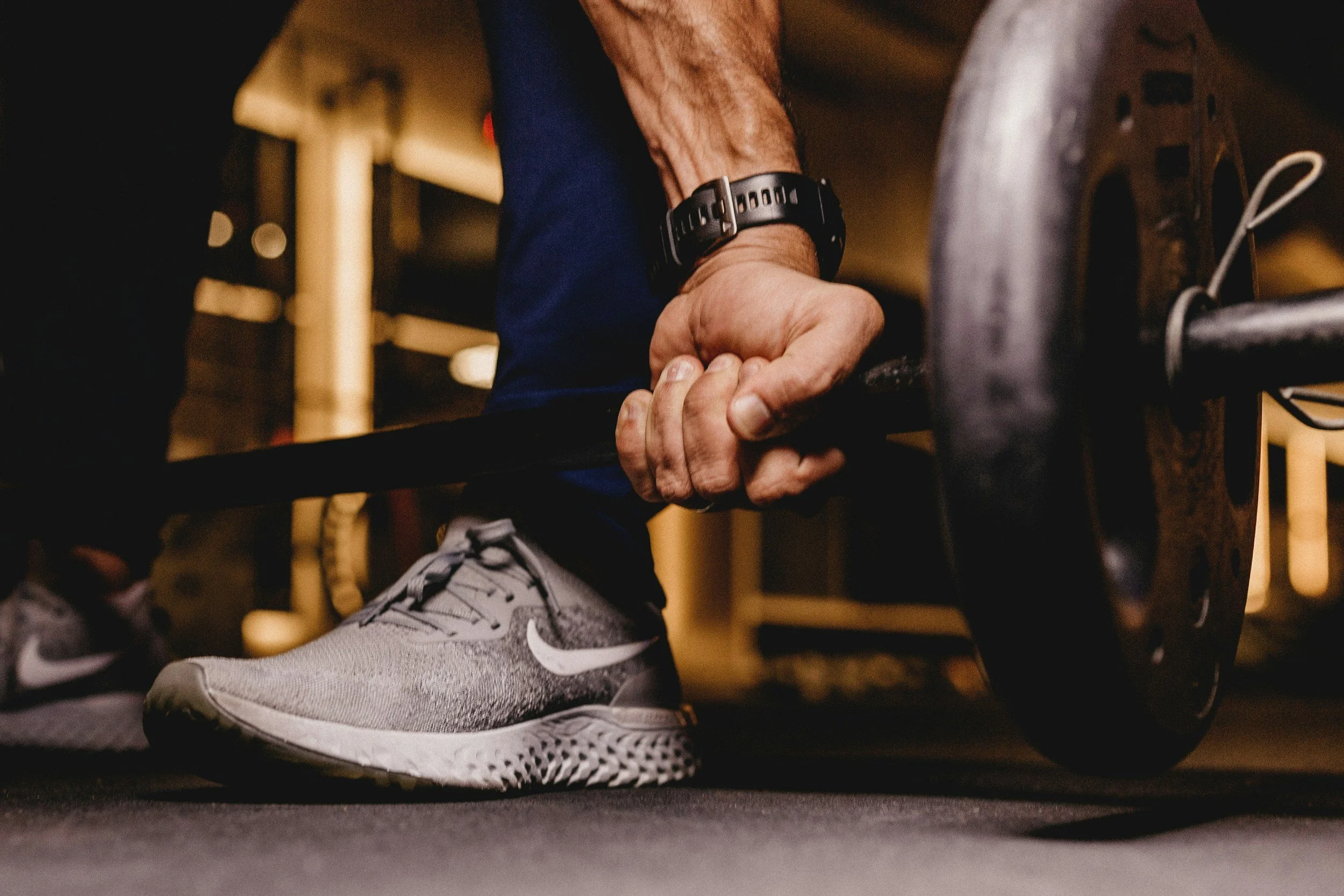 Close-up of a person's hand gripping a dumbbell while exercising in a gym, wearing a black watch and gray athletic shoes.