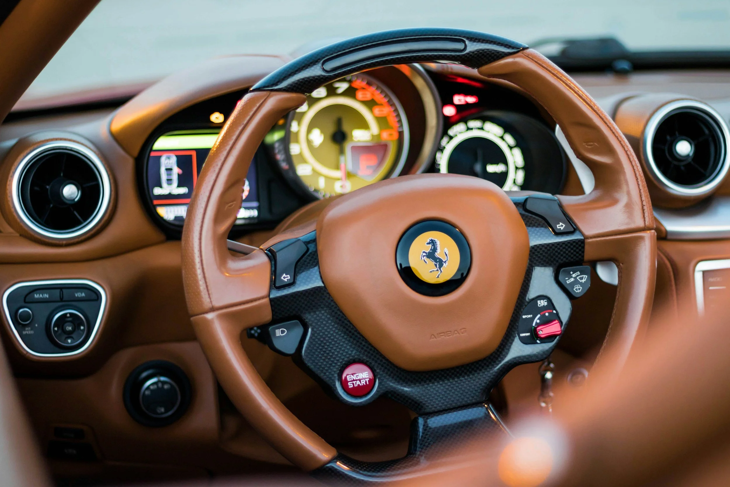 Interior view of a luxury car dashboard, featuring a brown leather steering wheel with a Ferrari logo, digital instrument cluster, and various control buttons.