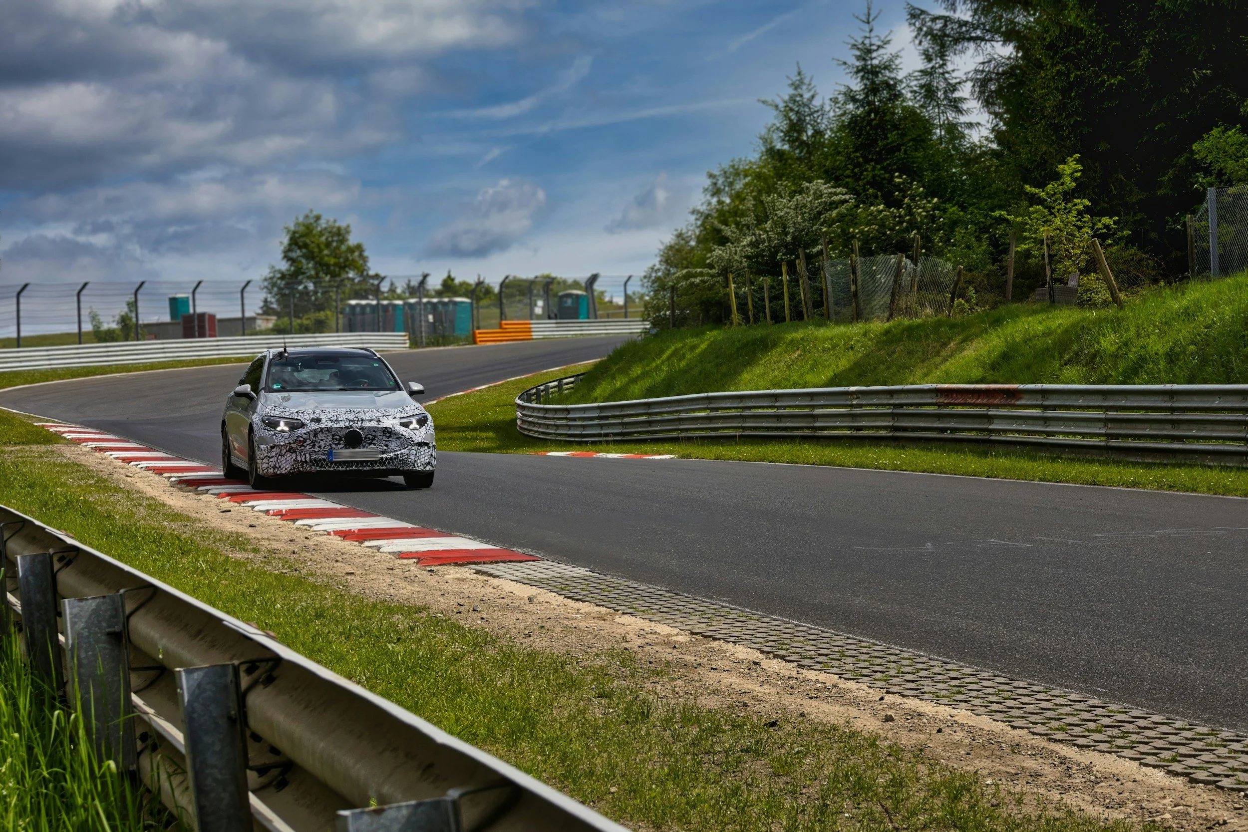 A silver test vehicle on a race track with red and white curb, surrounded by green grass, trees, and fencing, under a partly cloudy sky.