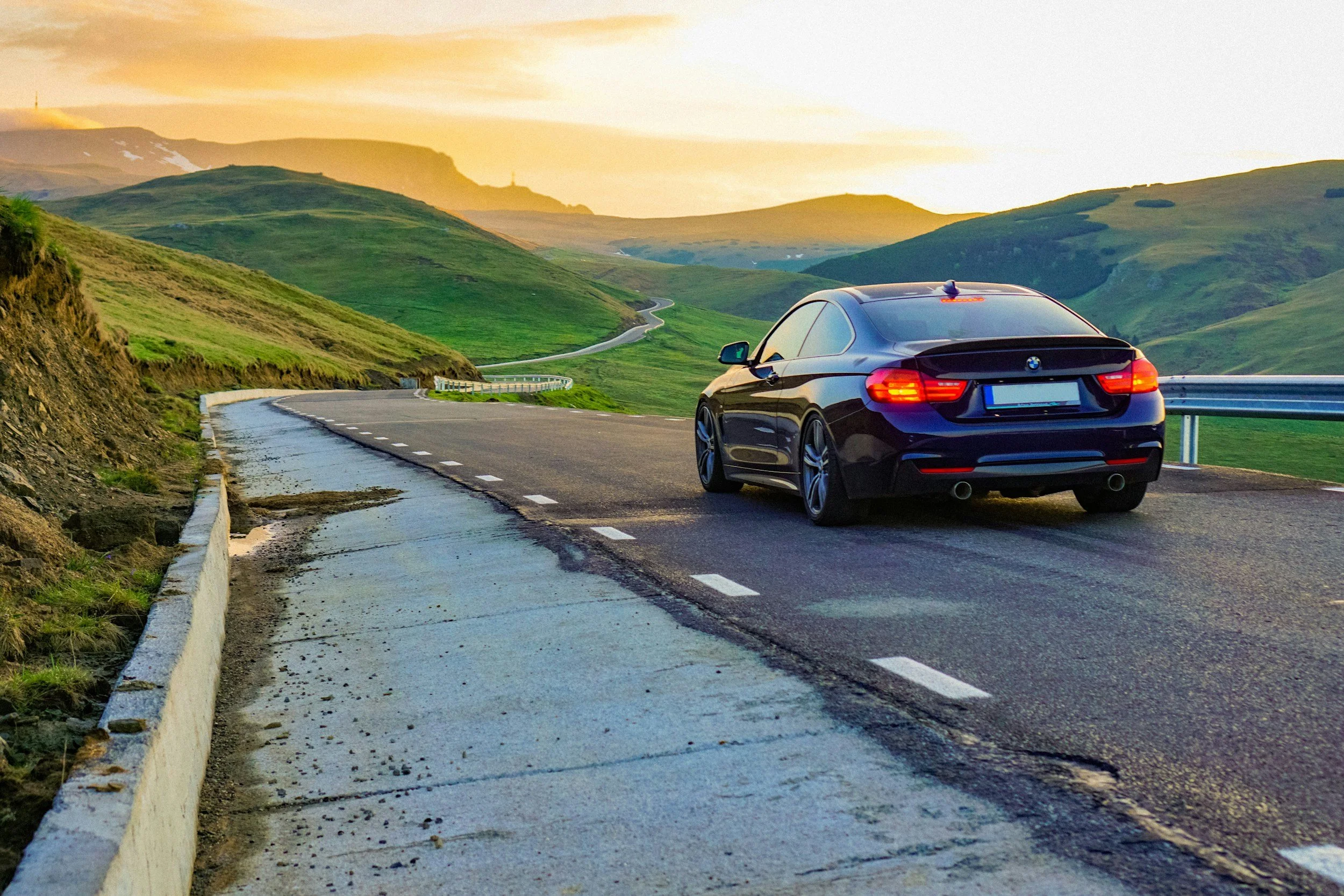 Black BMW parked on the side of a winding mountain road with green hills and sunset in the background.