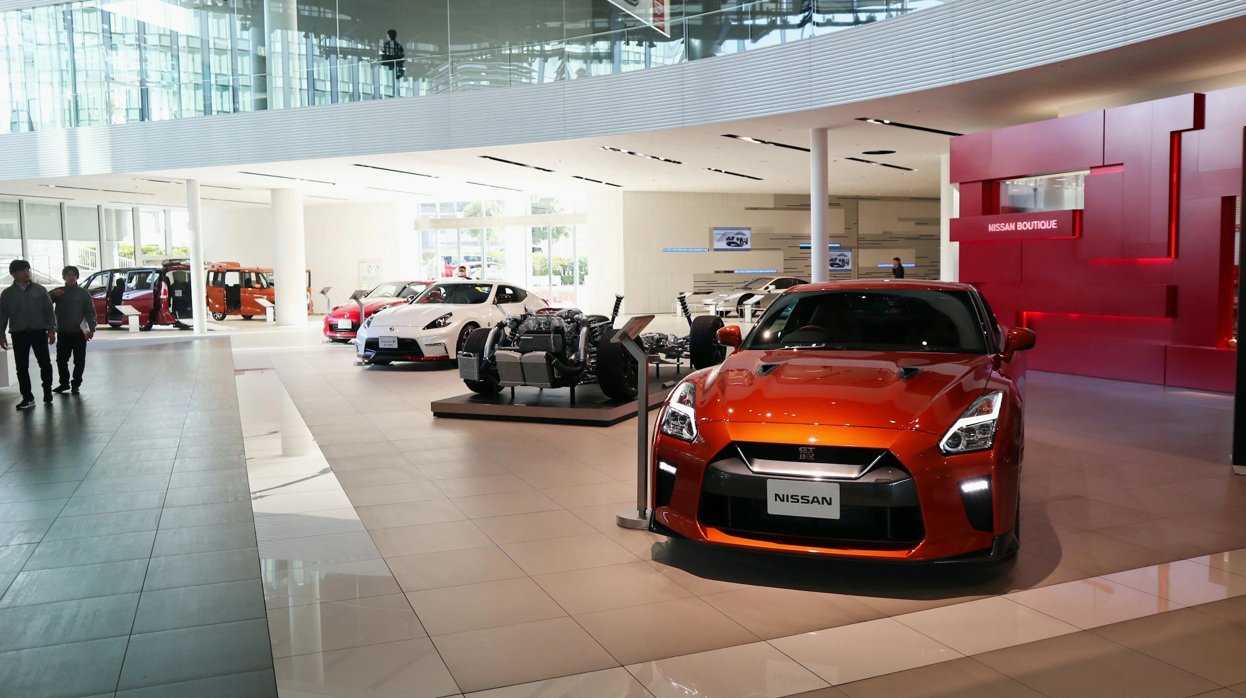 Interior of a Nissan car showroom with various Nissan vehicles on display, including a prominent red Nissan GT-R in the foreground, a white sports car, and some concept models, with a modern architectural design and natural light.