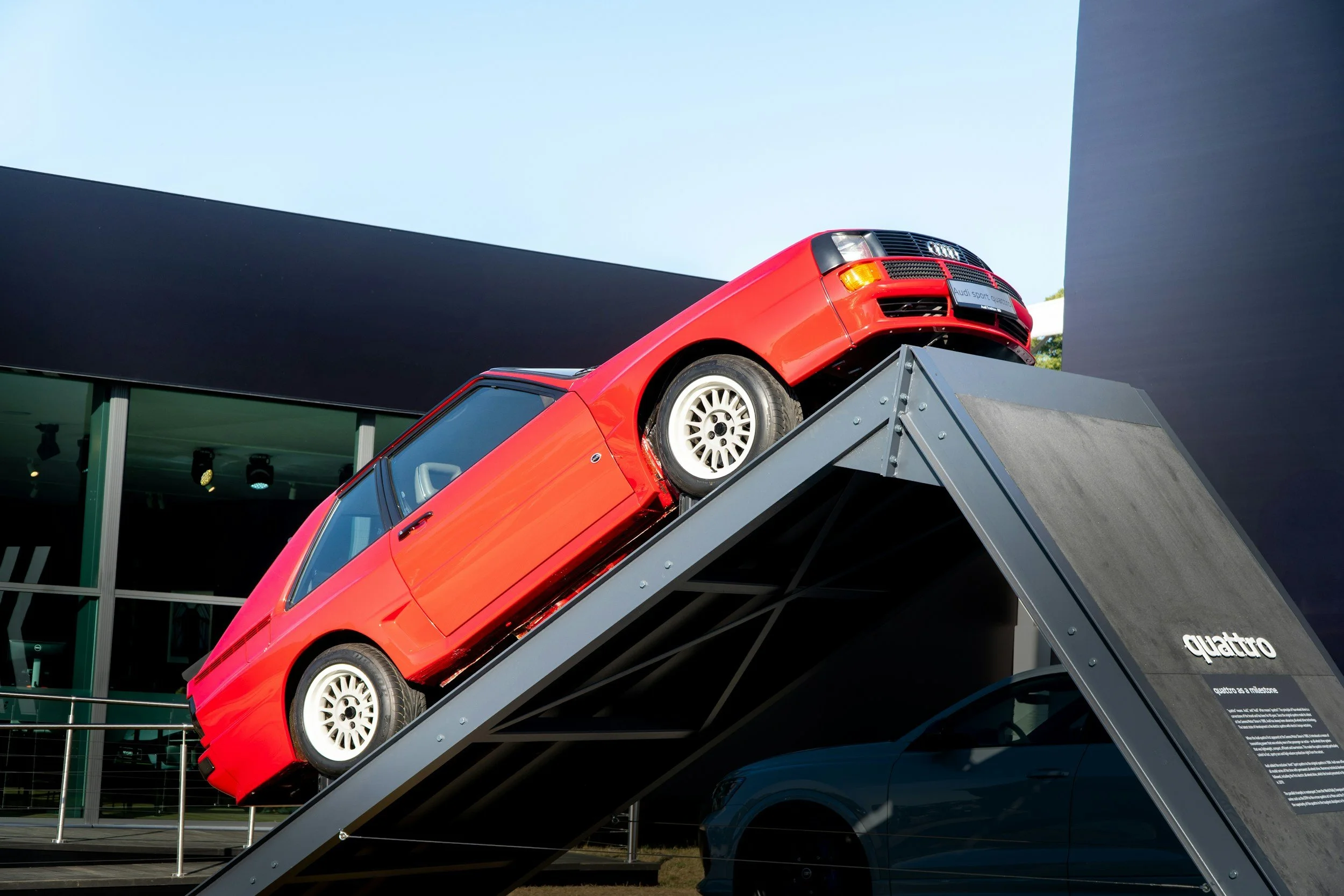 A red race car on display at an auto show, mounted on a tilted platform with a black front grille and white wheels, with black and gray exhibition structures in the background.