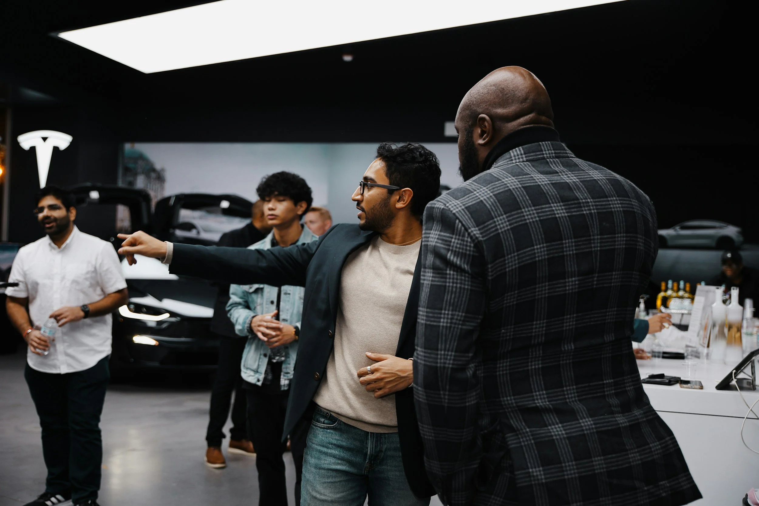 A group of diverse men gathered in a Tesla store, with cars on display and a sales counter in the background, engaging in conversation.
