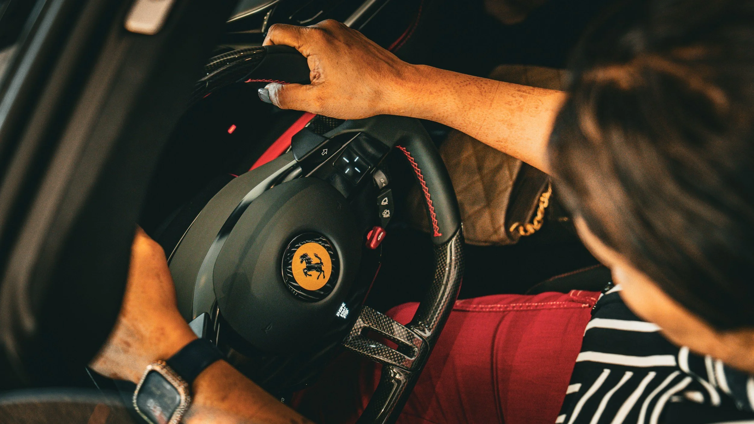 Person gripping a steering wheel with Ferrari logo, wearing red pants and a watch, inside a car.