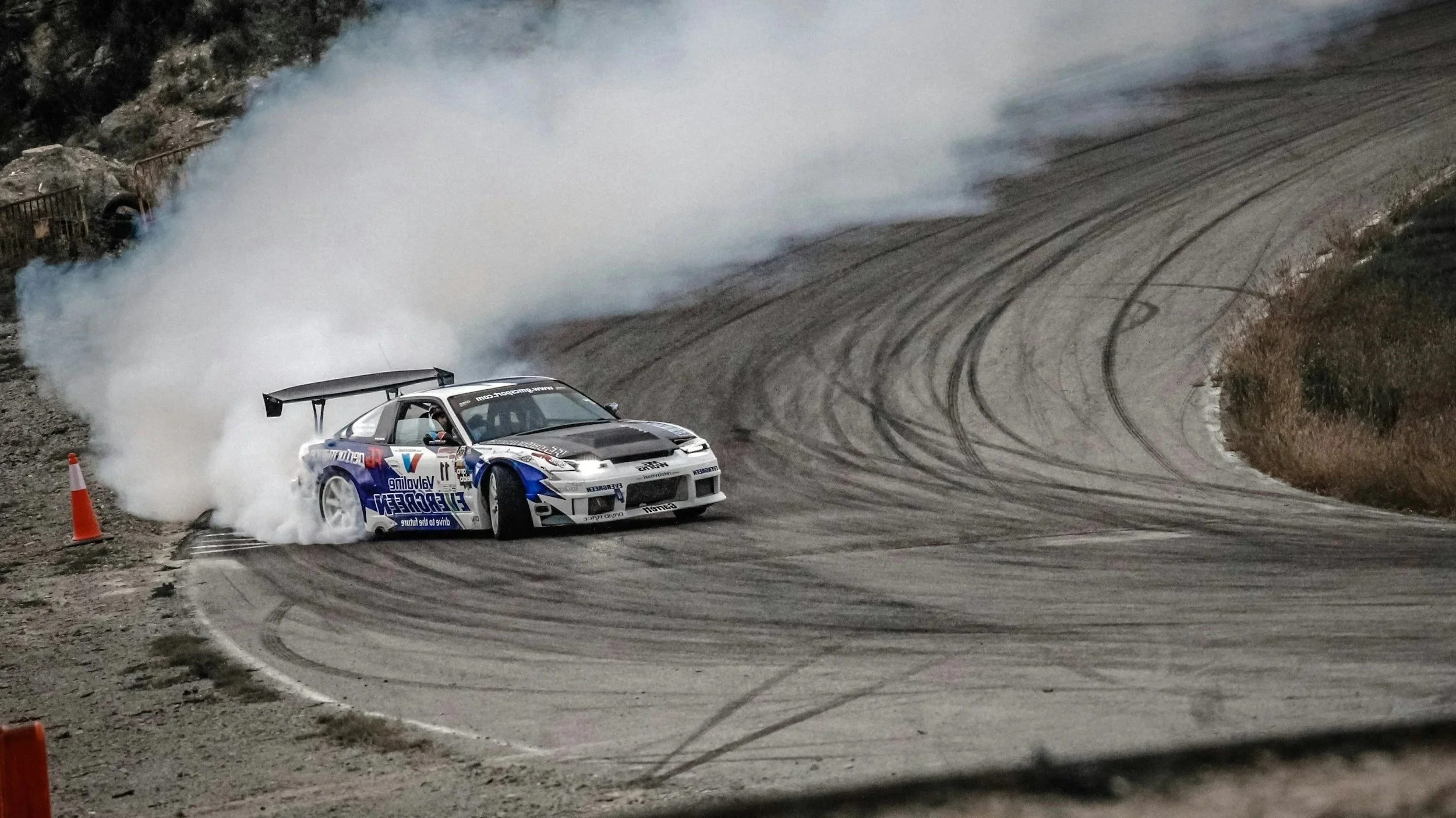 A black BMW M5 Drifting at Silverstone, with smoke coming from the rear tires and blurred background showing a race facility and trees.