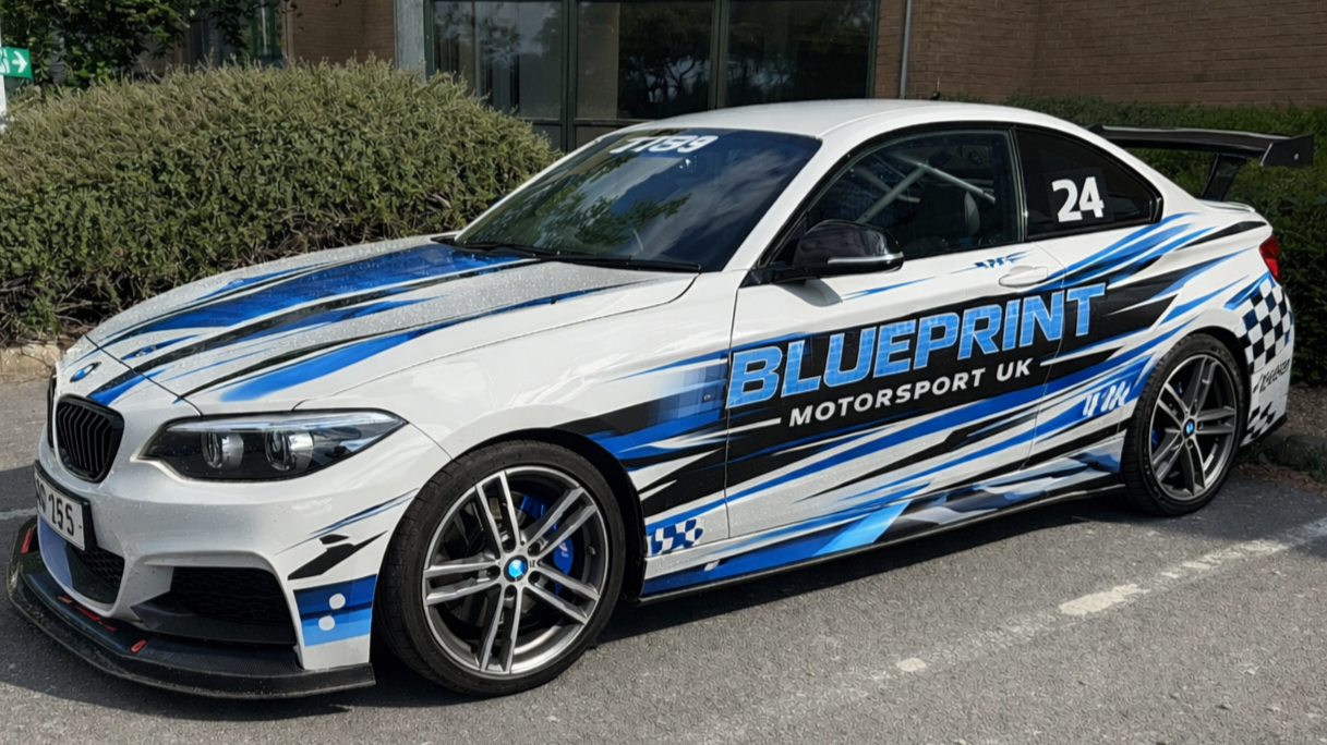 White BMW M240i race car with blue and black graphics, a large rear spoiler, and the words 'BLUEPRINT Motorsport UK' on the side.