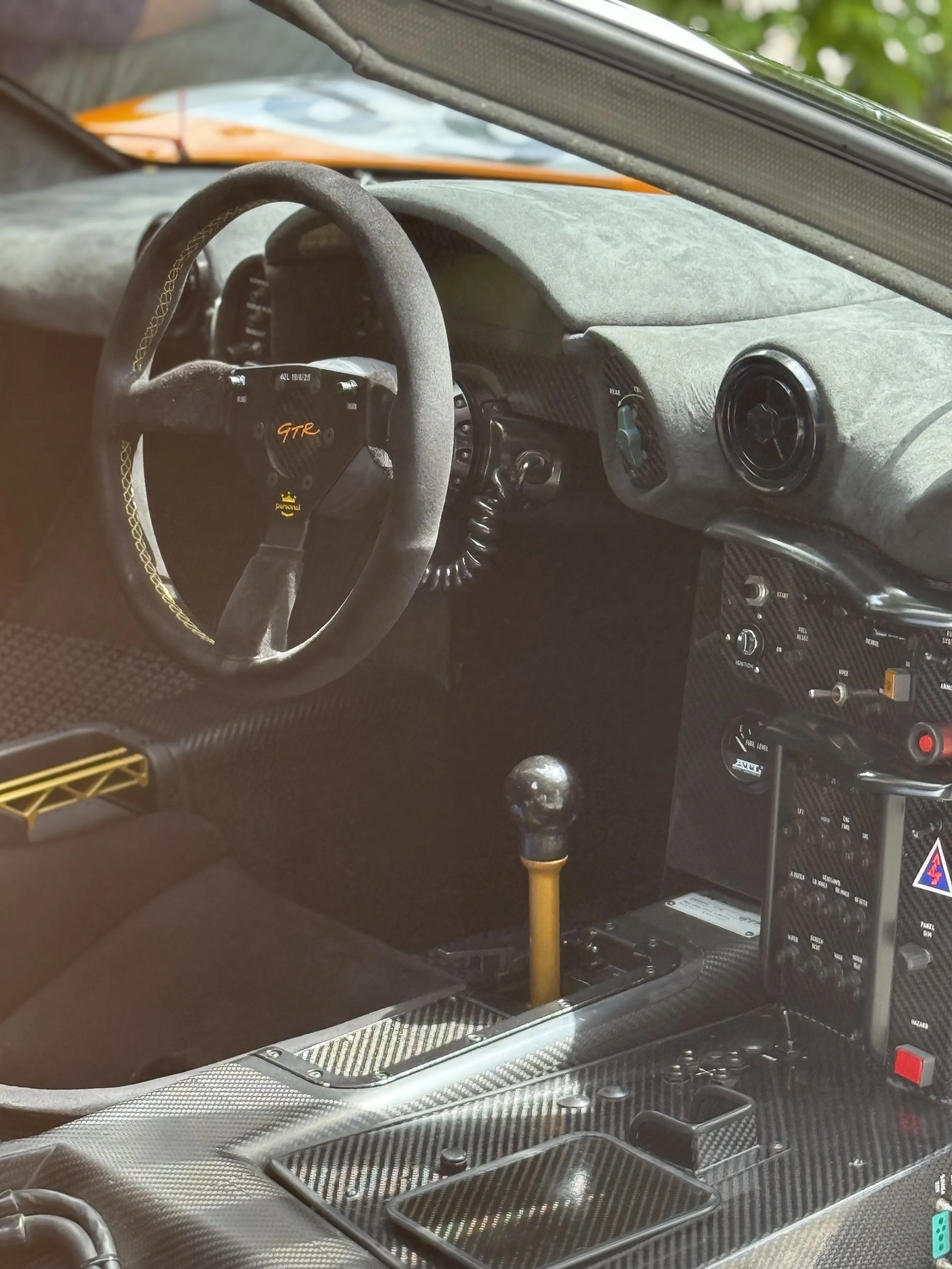 Interior of a race car with a racing steering wheel, carbon fiber dashboard, yellow gear shift, and various control switches.