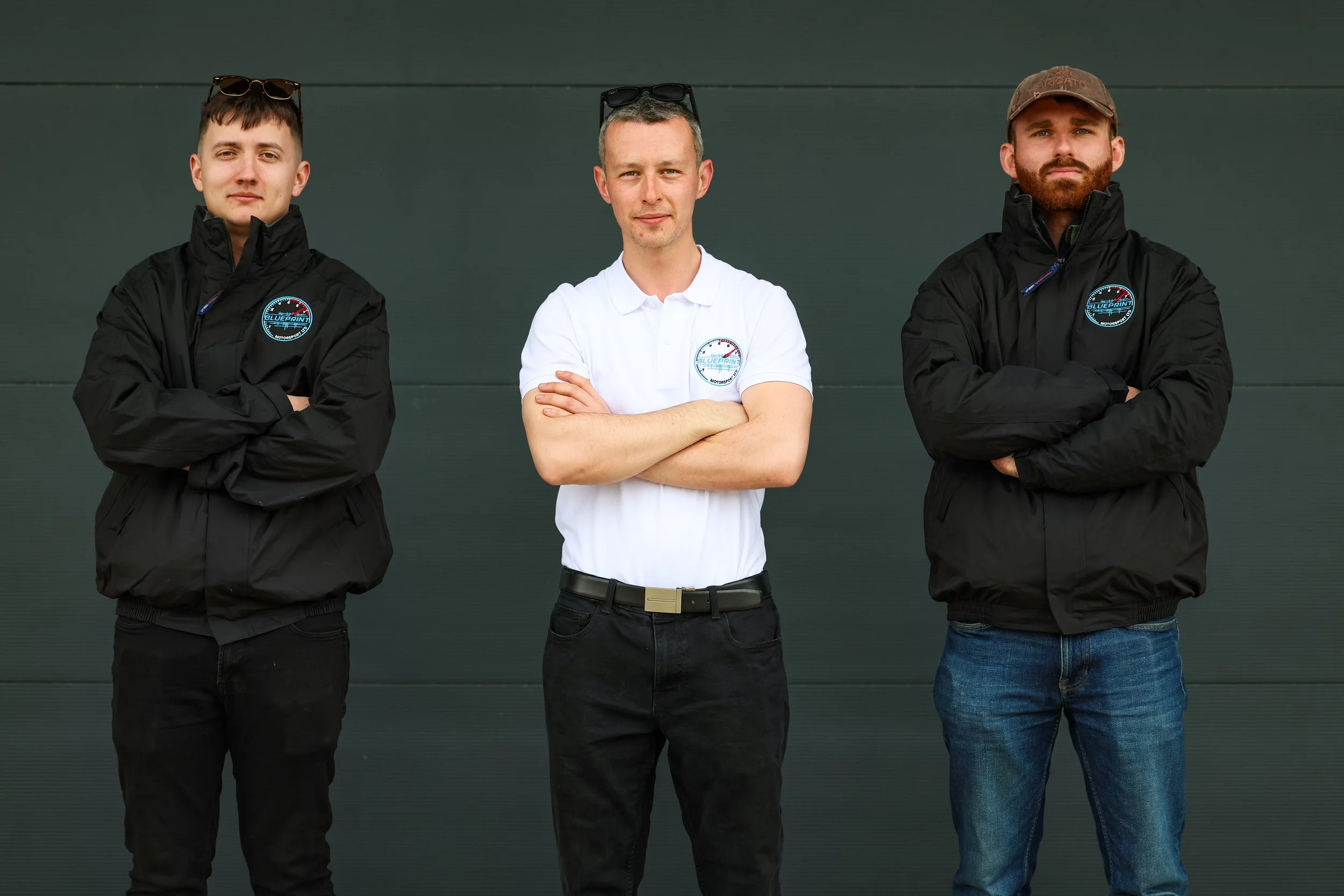 Three men standing with arms crossed in front of a dark green wall, wearing jackets and casual clothing, with sunglasses on their heads.