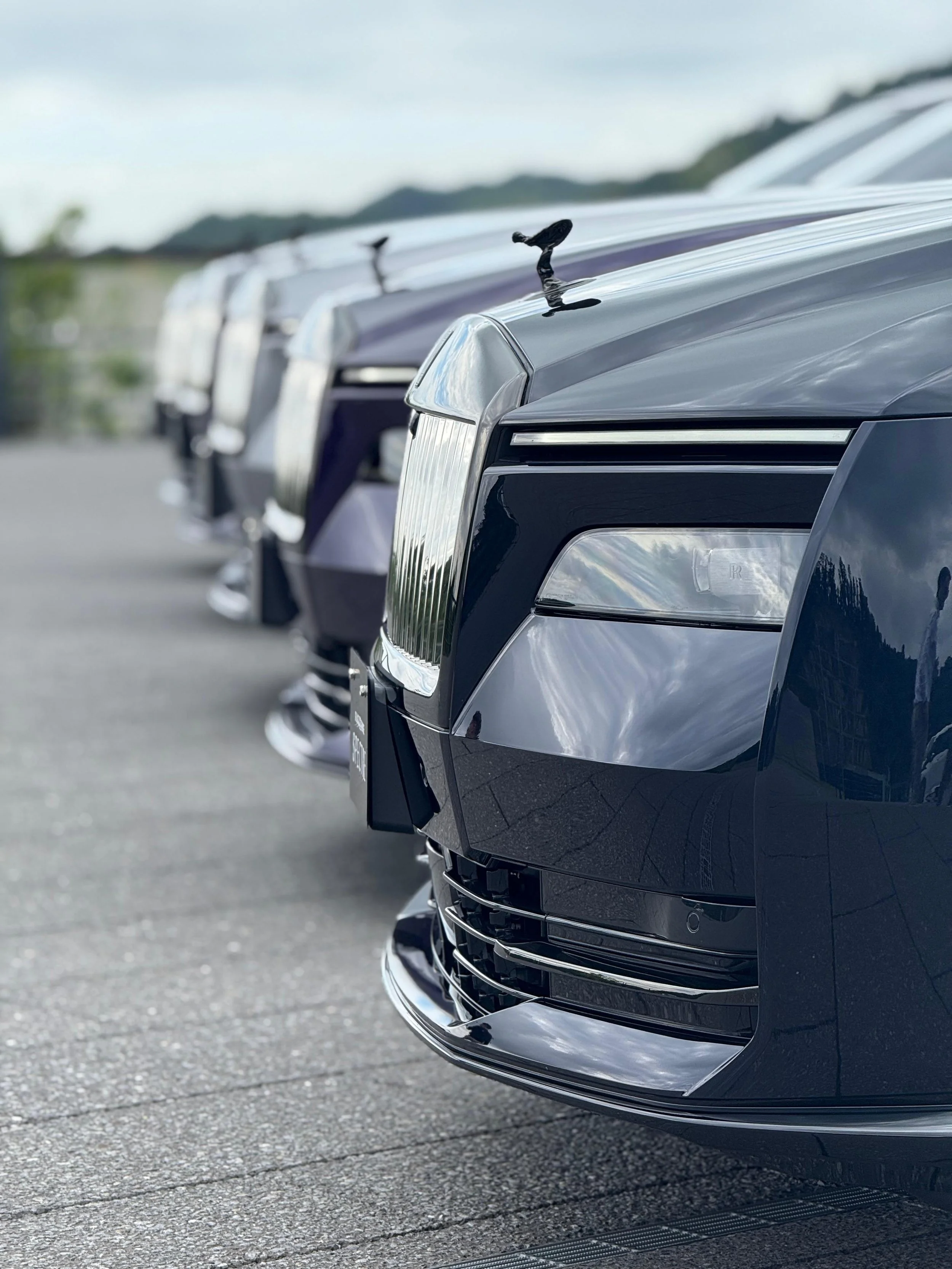 Close-up of the front of a row of new, shiny black luxury cars parked on a road, with mountains in the background.