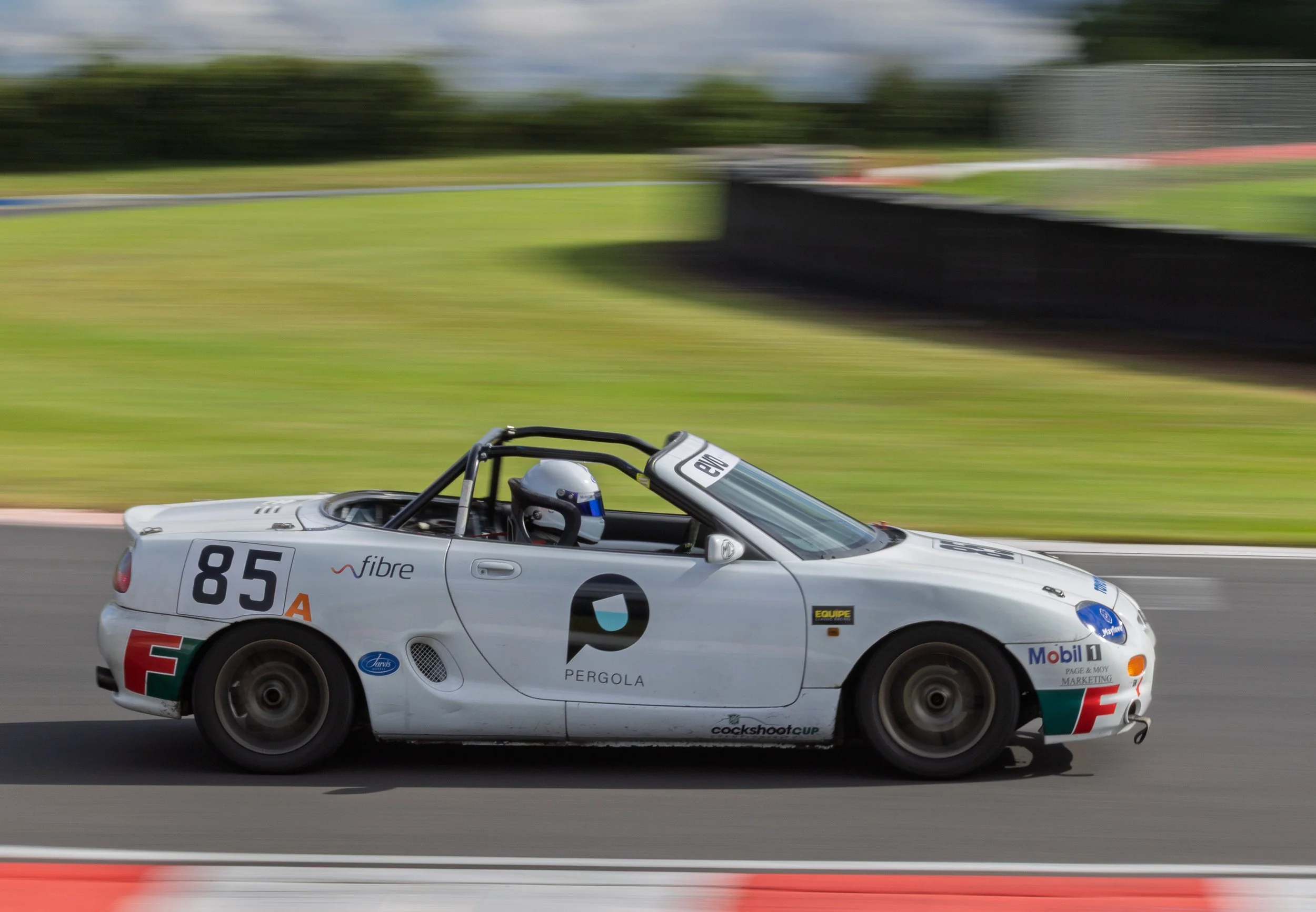 A white race car with the number 85 speeding on a racetrack, driver wearing a helmet inside the car, with blurred background indicating high speed.