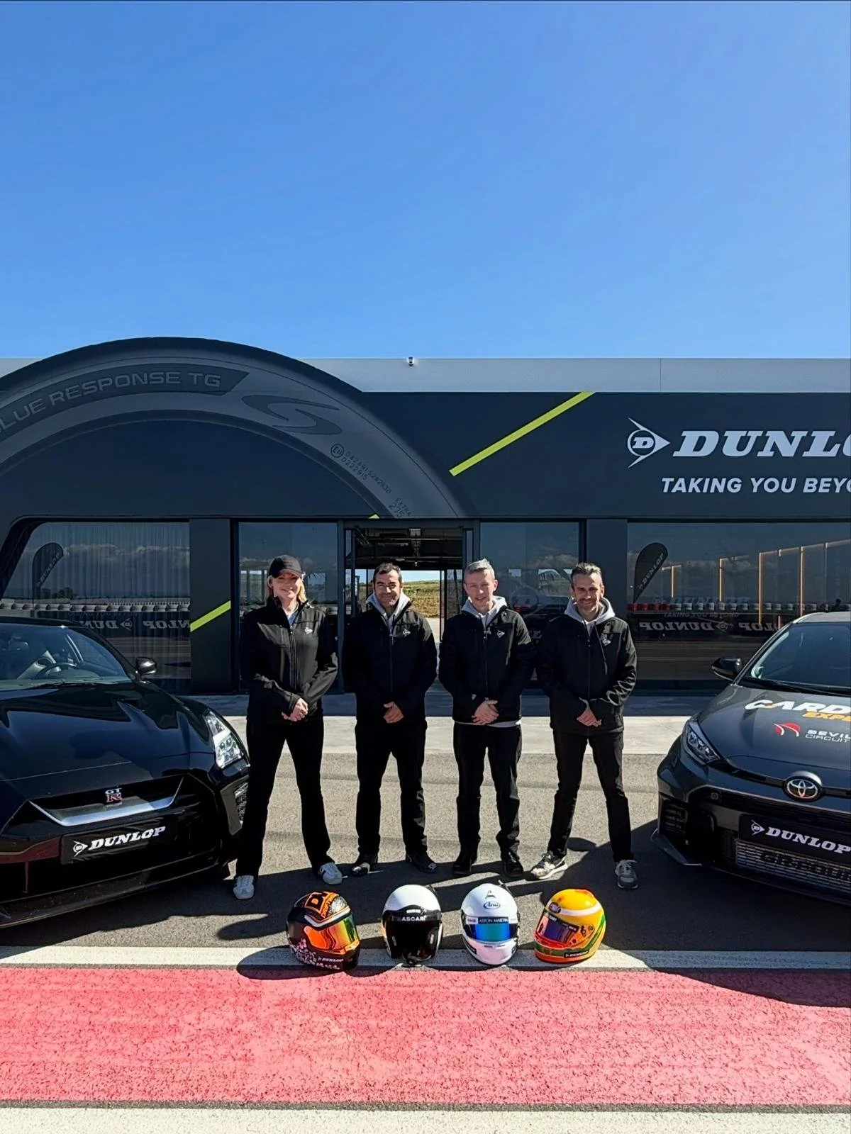 Four people stand in front of a Dunlop branded building with two race cars, race helmets on the ground, and clear blue sky.