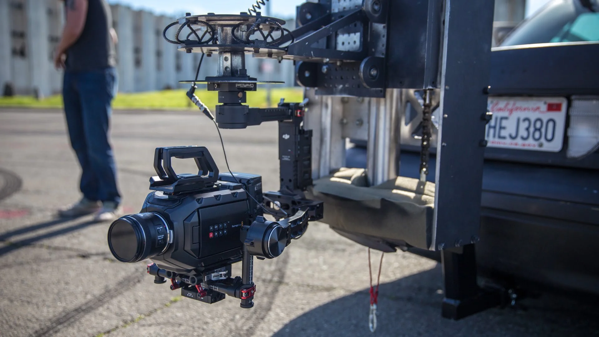 Close-up of a professional camera mounted on a mechanical rig attached to the back of a vehicle, with a person standing in the background.