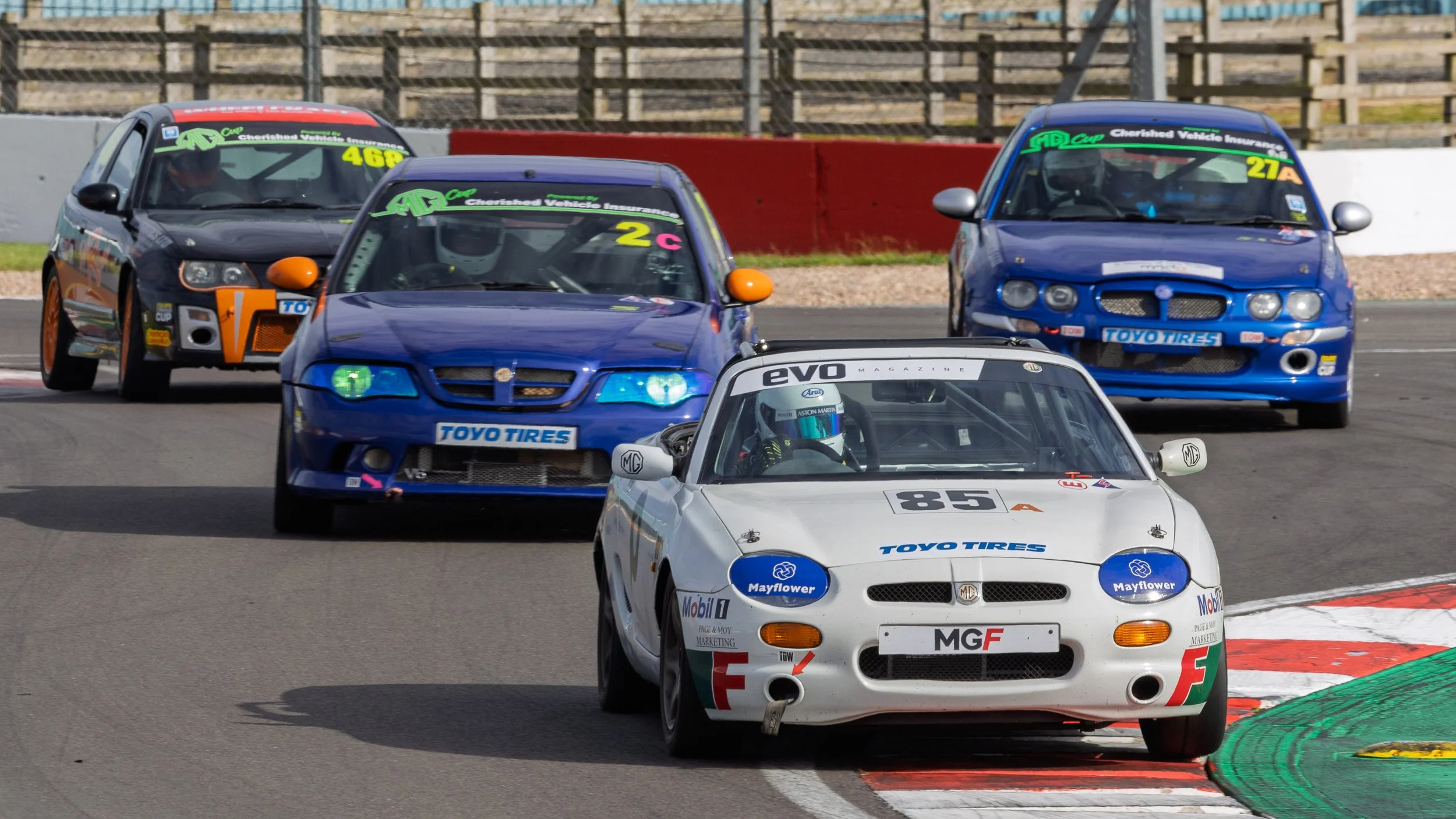 Multiple race cars navigating a turn on a racetrack, with a white MG race car in front, followed by blue and black cars.