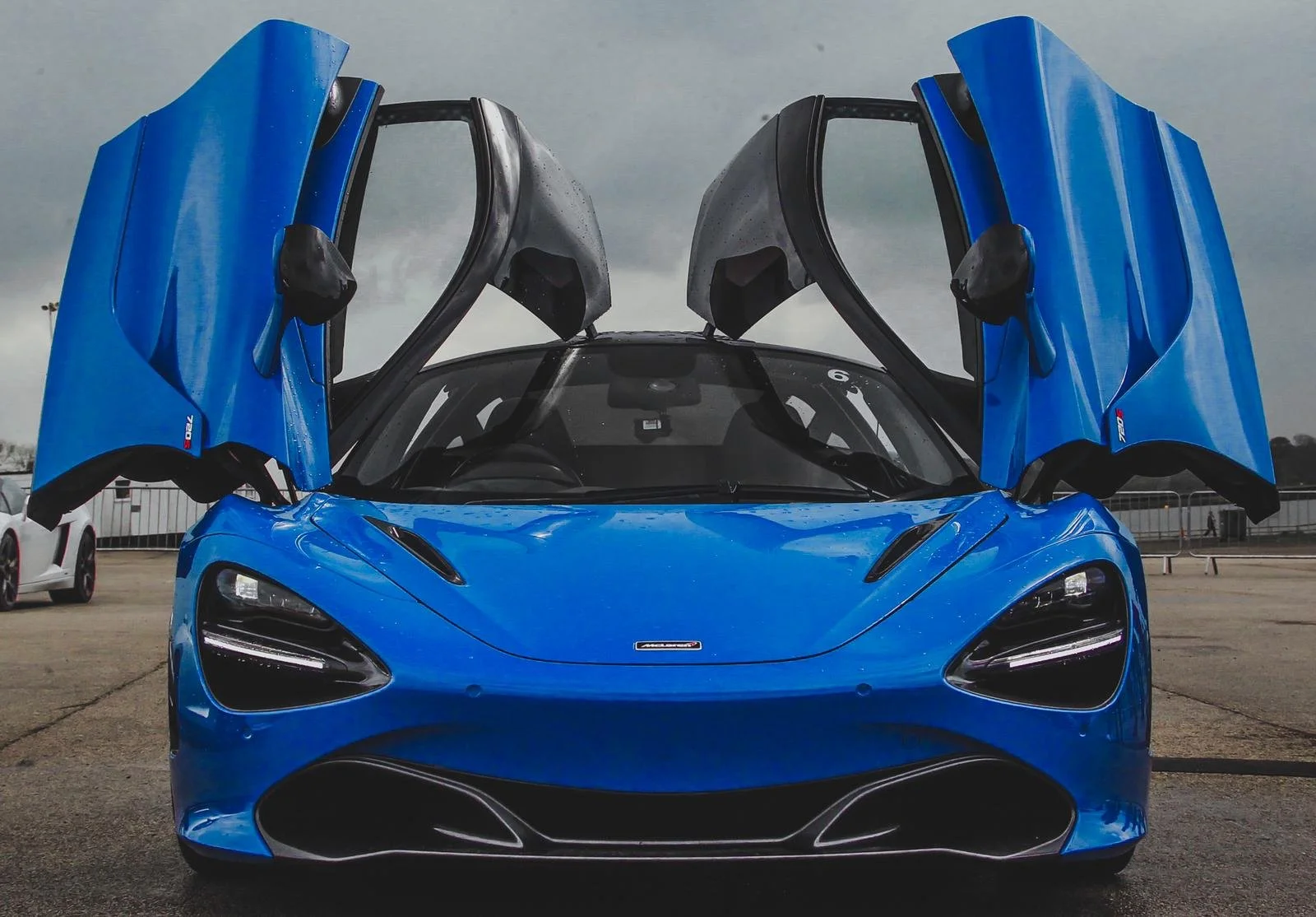 A bright blue McLaren sports car with its butterfly doors open, parked outdoors on a concrete surface under a cloudy sky.