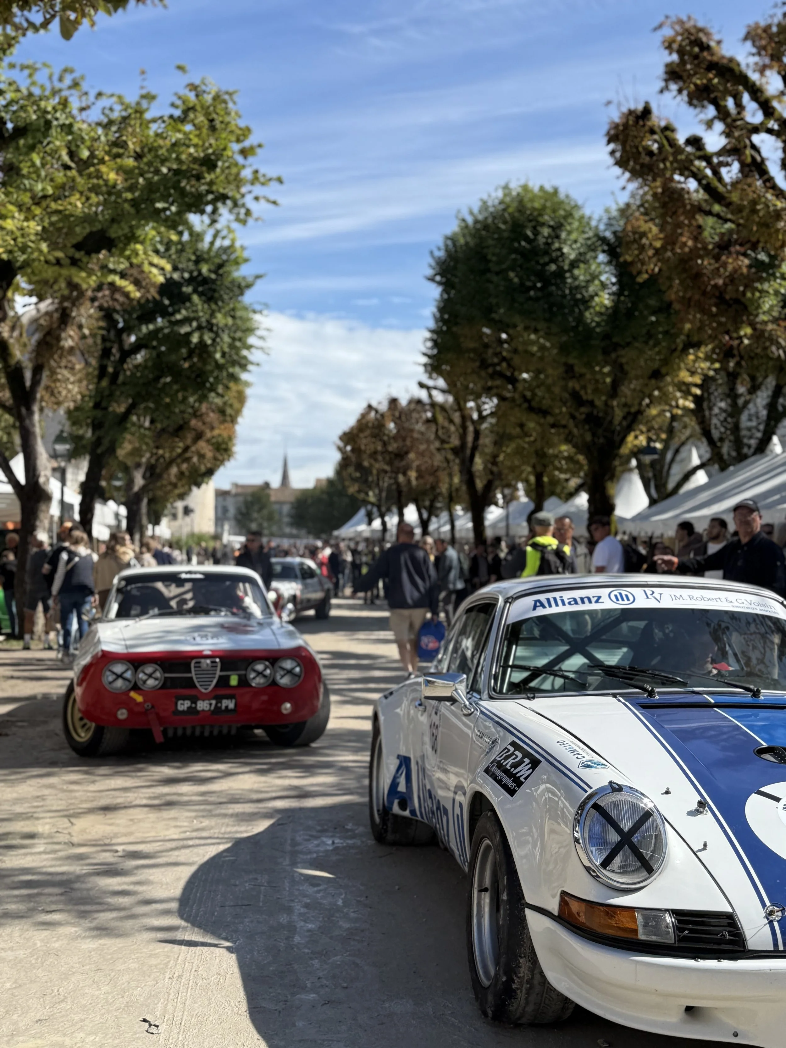 Classic race cars on display at an outdoor event with a crowd of people, tents, and trees under a partly cloudy sky.