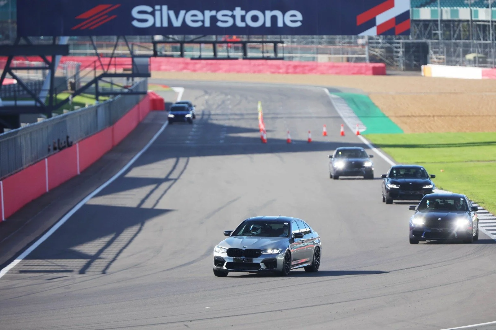 Multiple black and silver cars driving on a racetrack, with a Silverstone banner overhead and orange cones blocking part of the track.