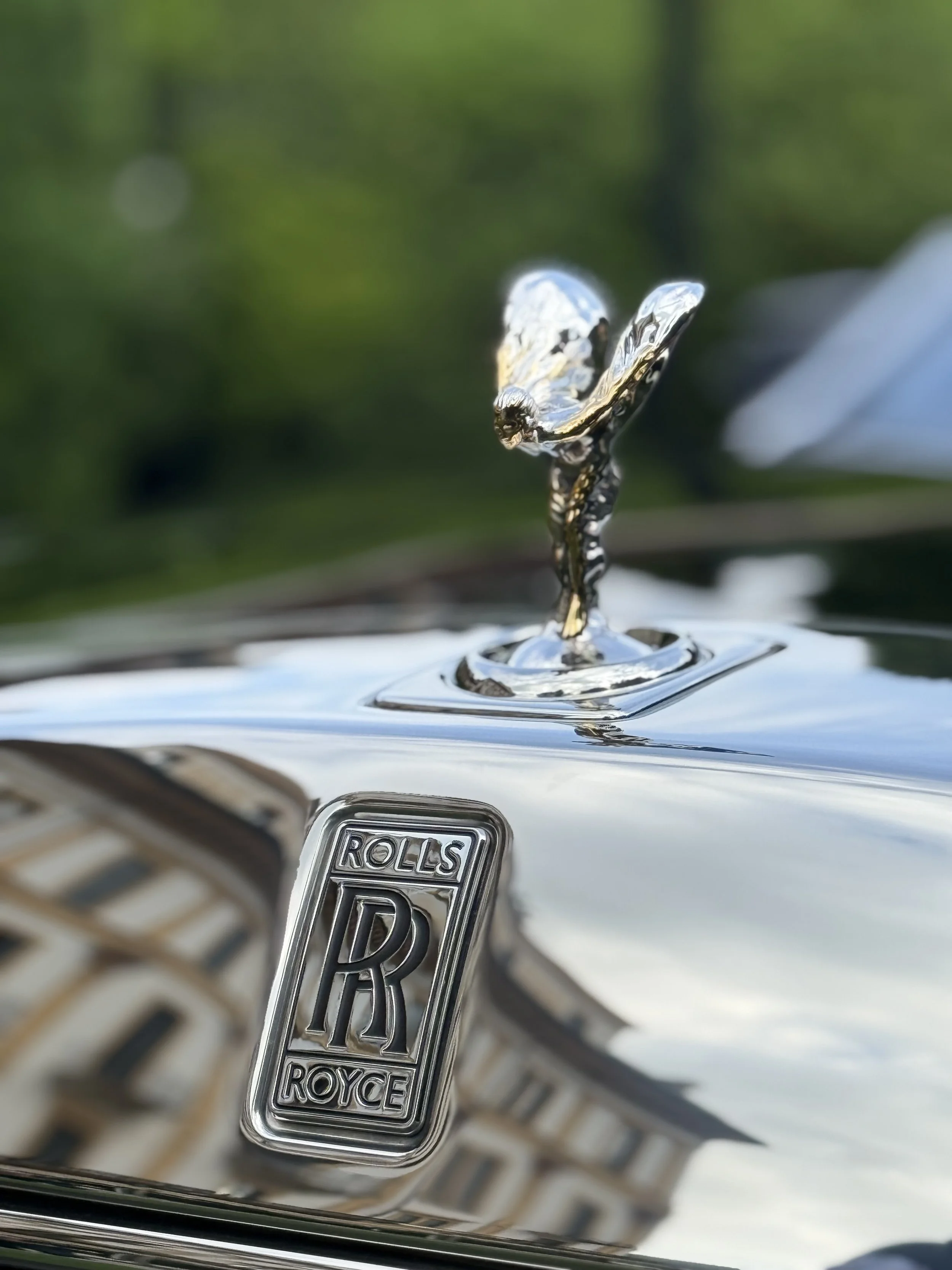 Close-up of a Rolls-Royce hood ornament and emblem on a luxury car with a building reflected on the chrome surface.