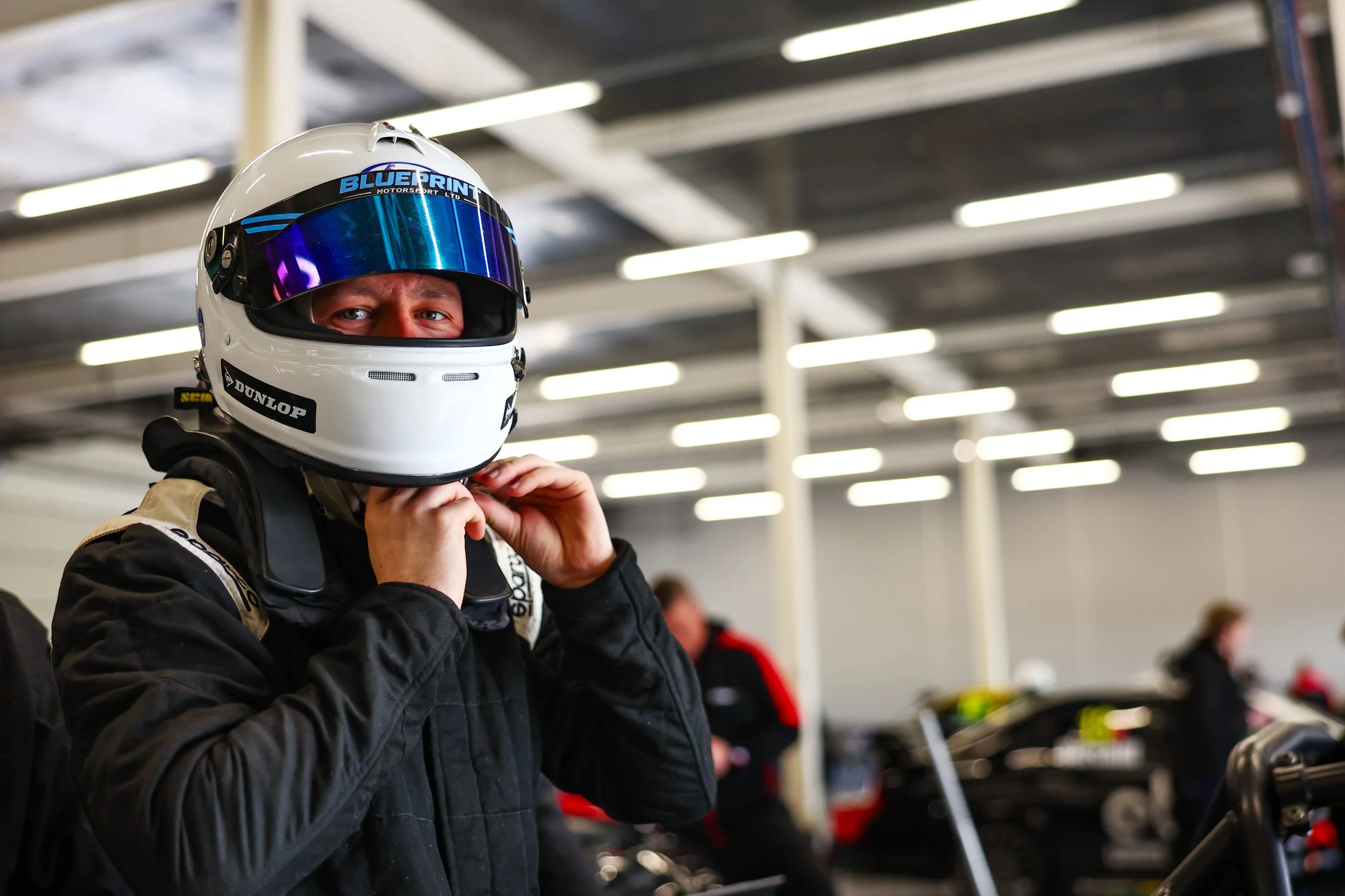 Race car driver wearing a white helmet with blue and black accents, and a black racing suit, in a garage or pit area.