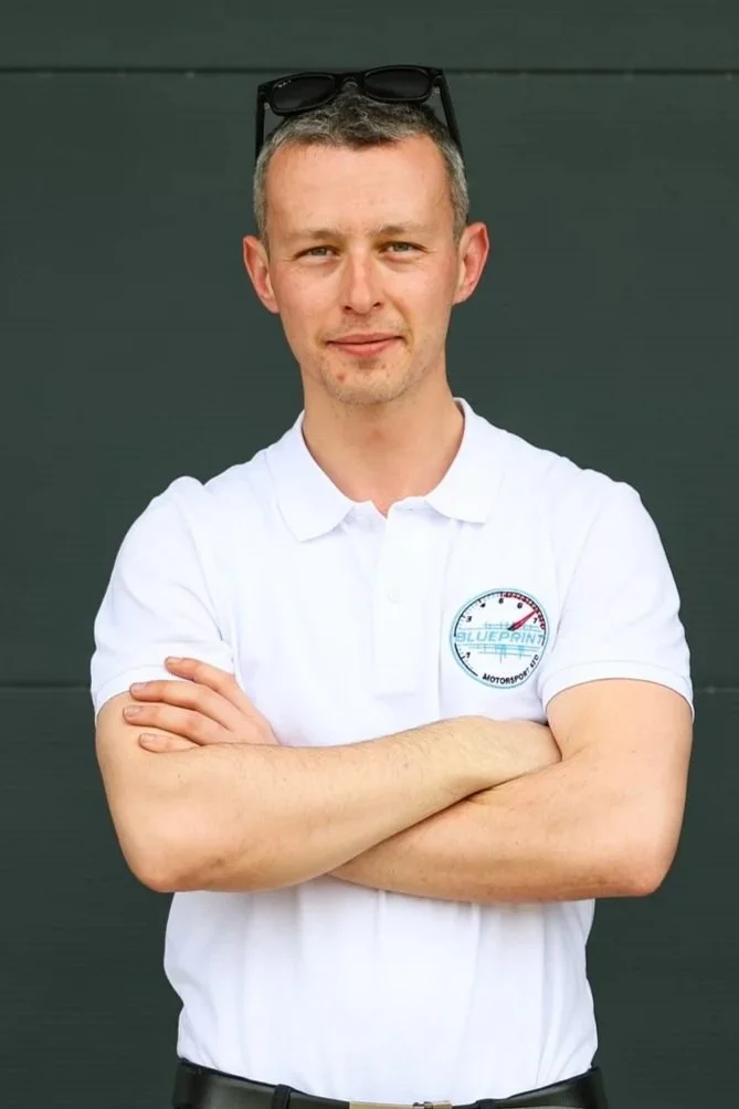 A man with short, gray hair and sunglasses resting on his head, wearing a white polo shirt with a logo, standing with arms crossed against a dark green background.