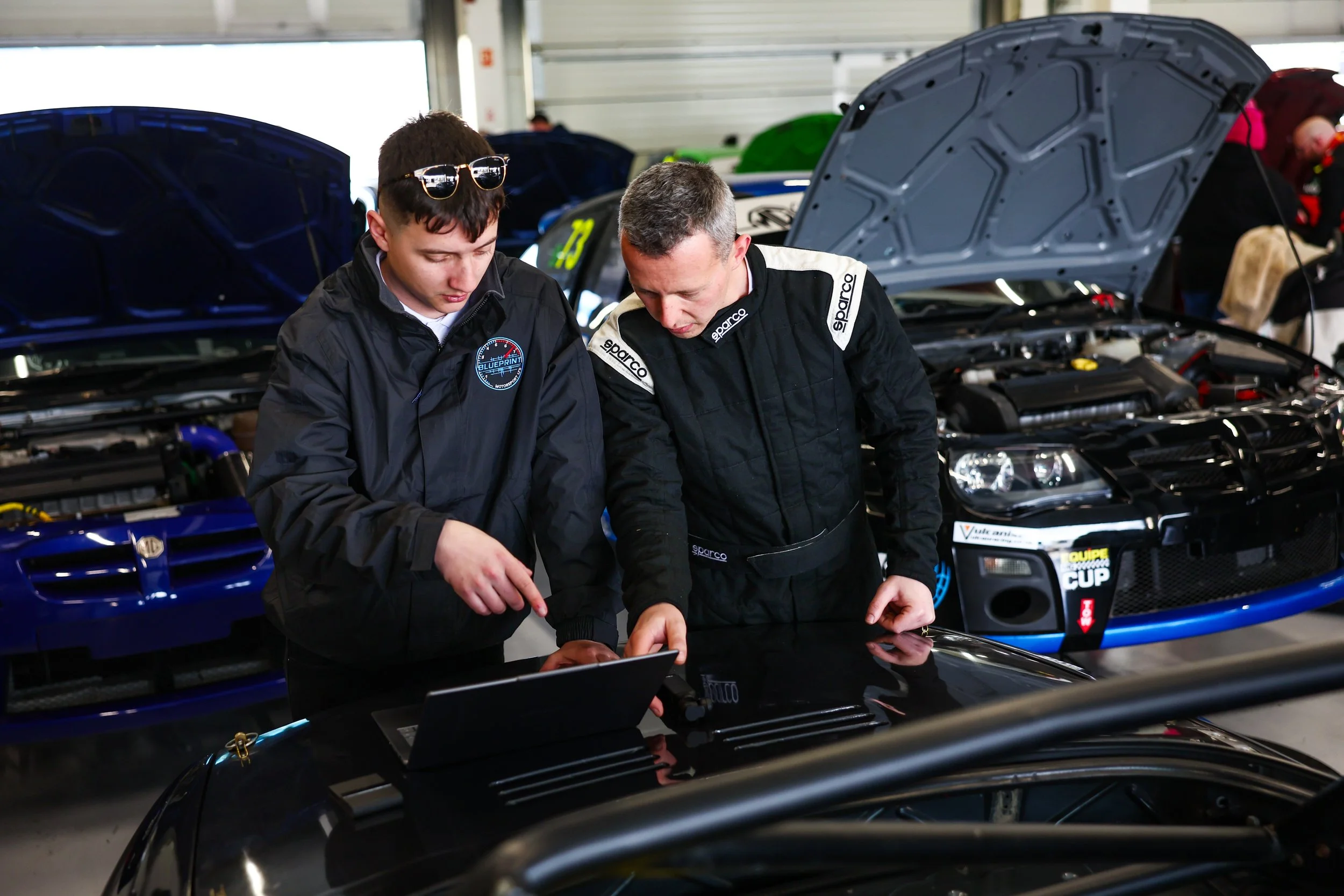 Two race car mechanics in black Sparco uniforms working together in a garage, with open hoods of race cars behind them.